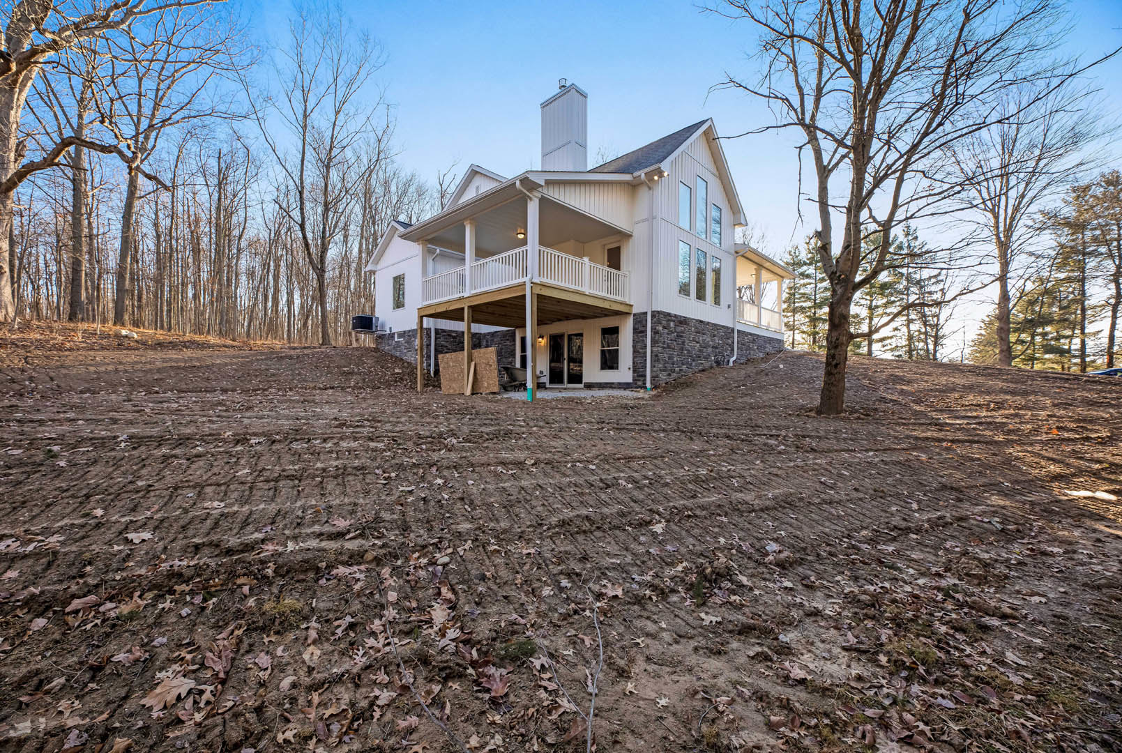 Two-story home with wide covered porch, white siding, brick chimney, large mature tree in front yard, multiple windows, and landscaped lawn