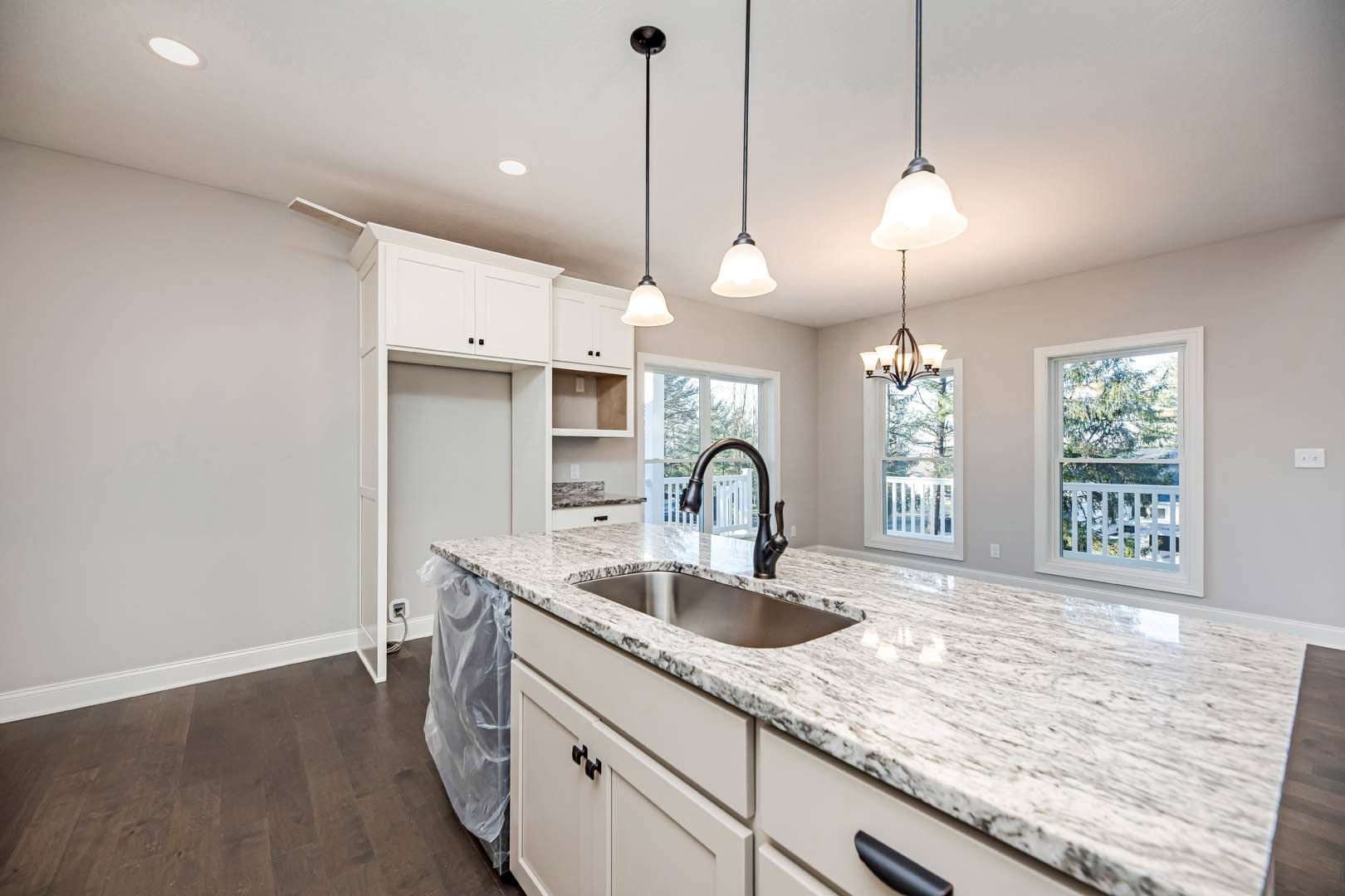 Bright kitchen with white cabinetry, stone countertop, stainless steel sink beneath a window with white railing, black pendant lamp overhead, and trees visible outside