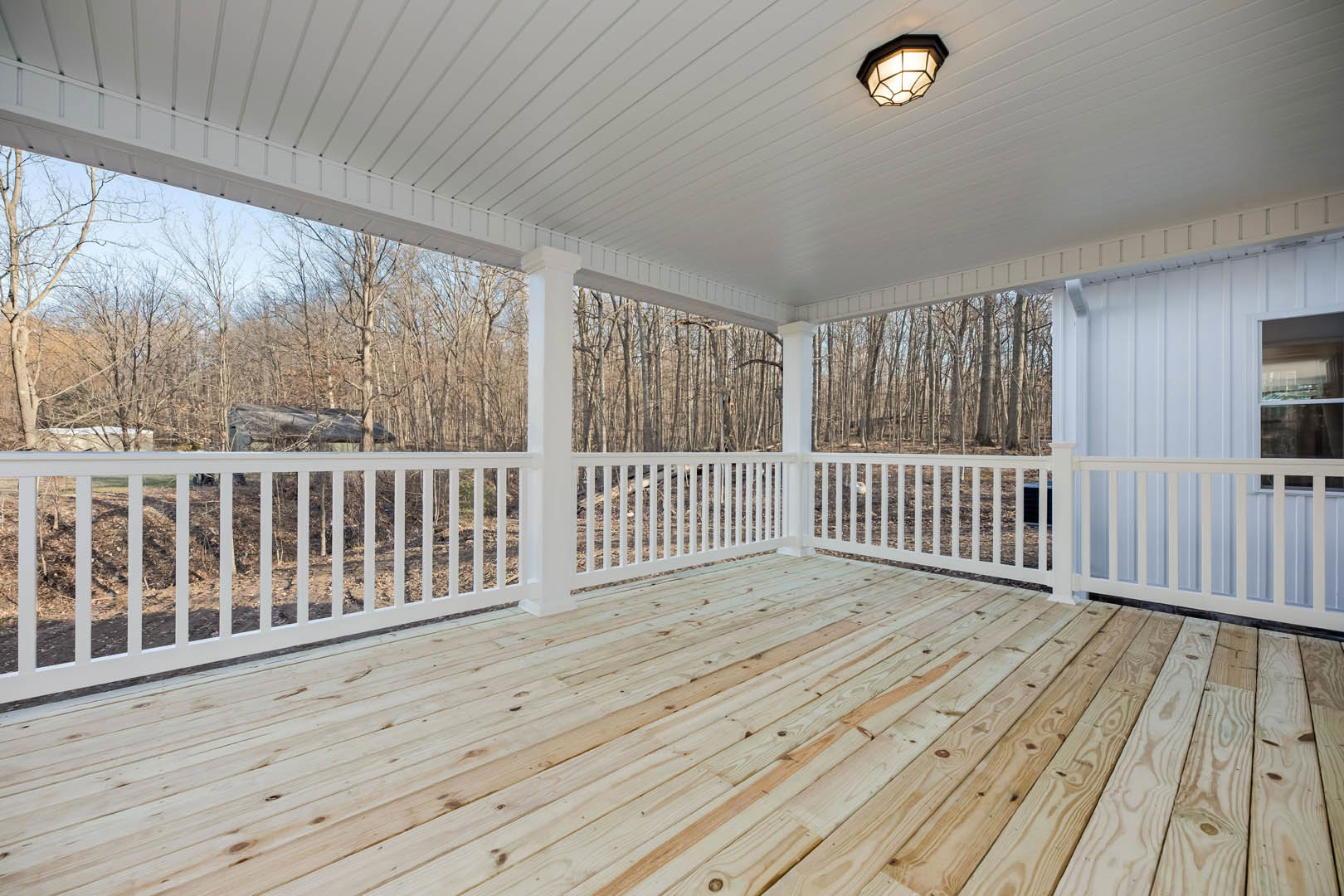Spacious deck with white railing and white-beamed ceiling, ceiling light fixture, overlooking leafless trees and wooded landscape