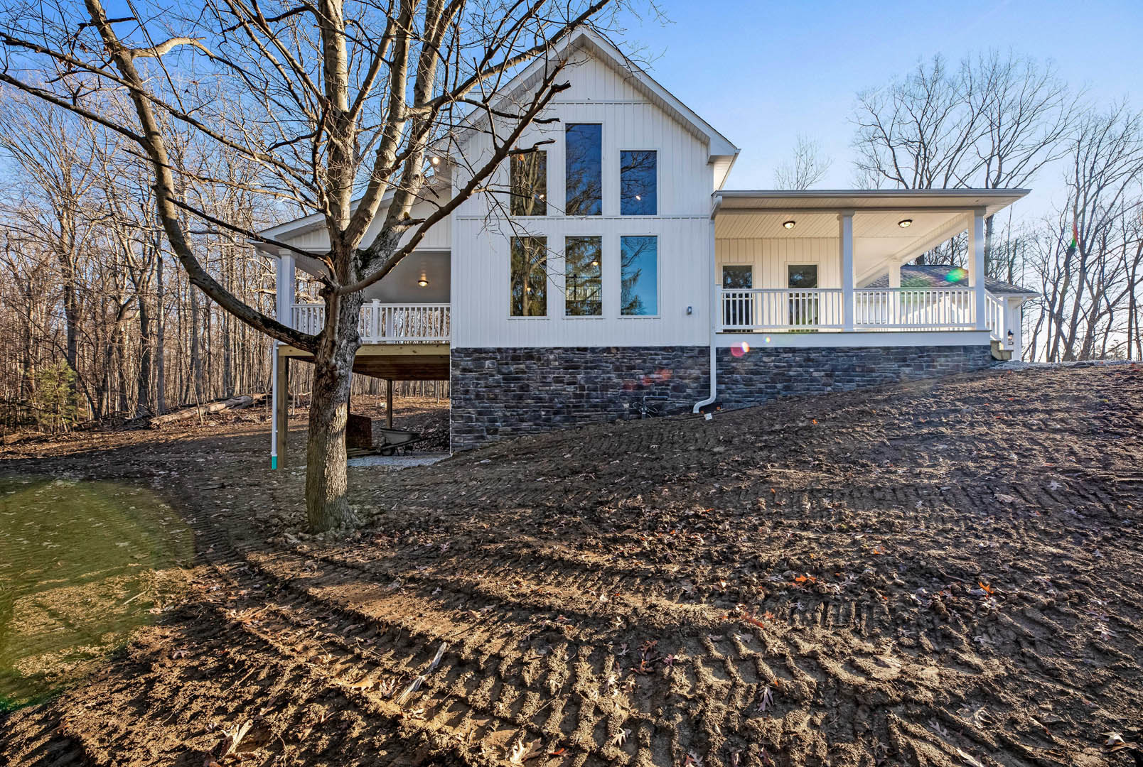 White house with large windows, tree with bare branches in backyard, dirt hill with tire tracks in foreground, utility pole with light, blue winter sky