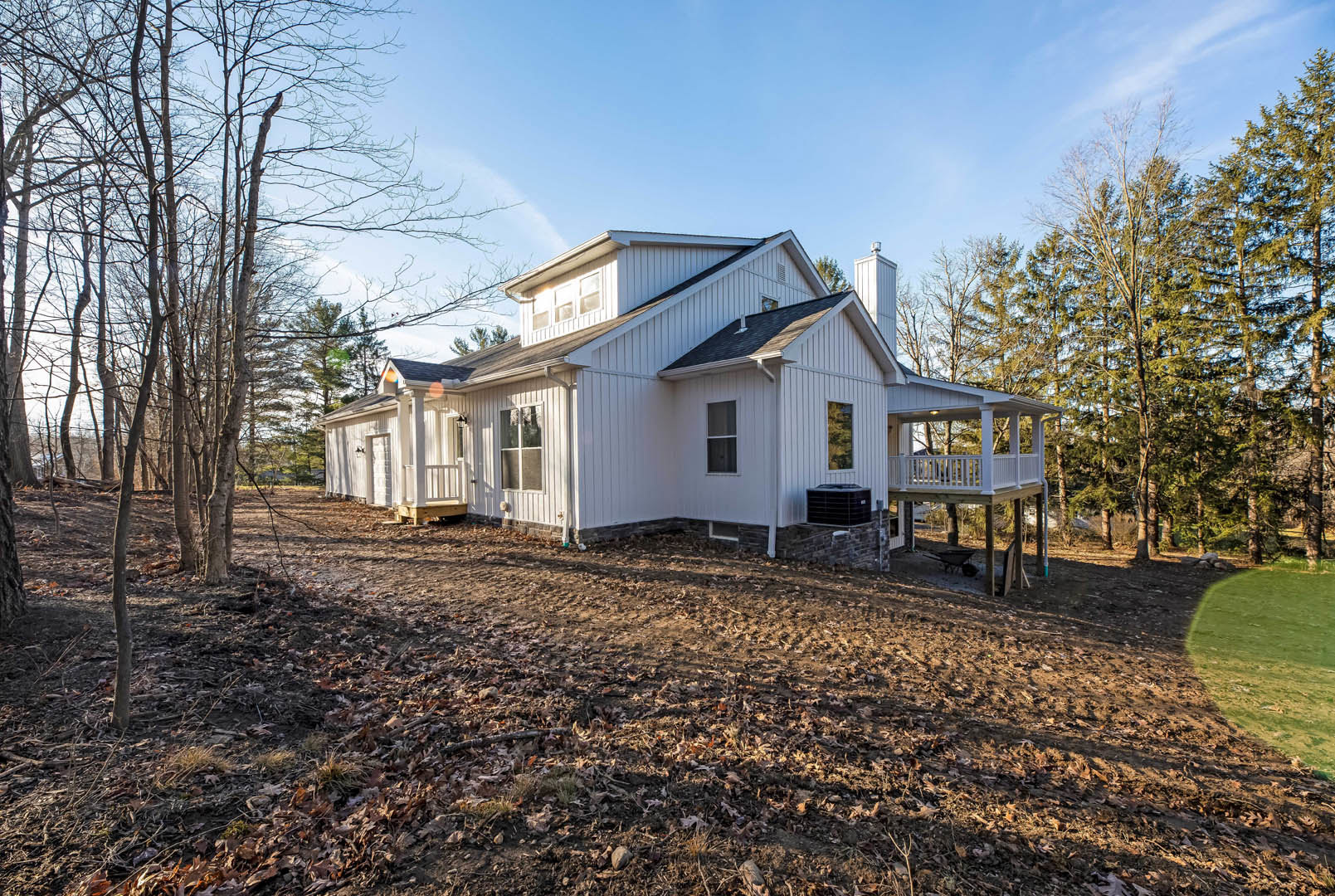 White house with black roof, elevated wooden deck, surrounded by mature trees, patchy green grass and dirt in front yard, large windows visible on exterior.