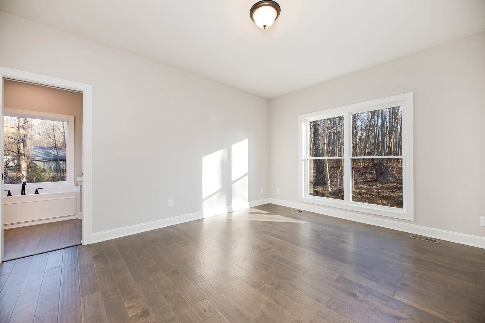 Spacious room with hardwood flooring, large windows framed in white, ceiling light fixture, and views of trees outside