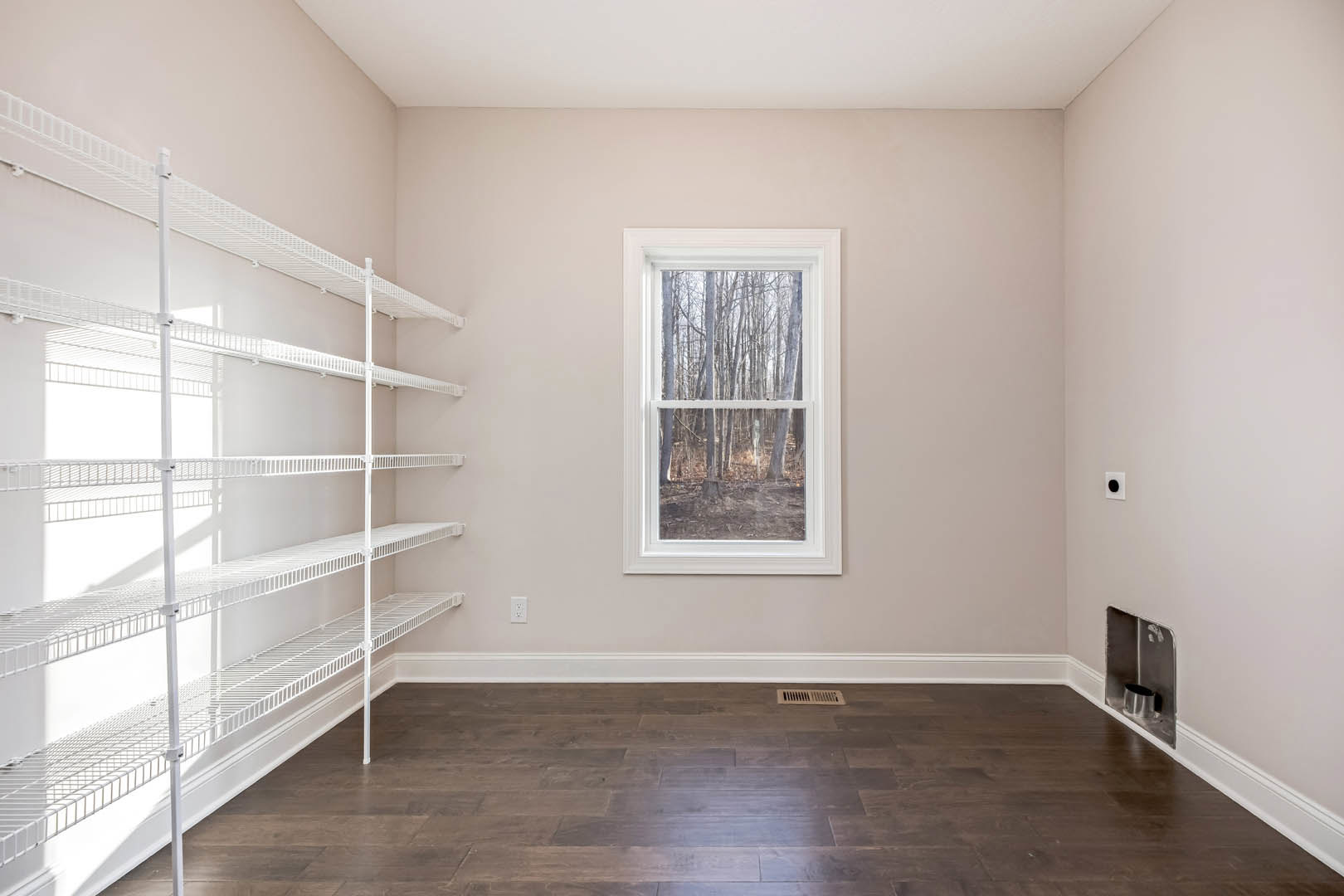 Empty room featuring built-in white shelves, wood laminate flooring with a floor vent, a window overlooking trees, and a silver cup placed on a shelf.