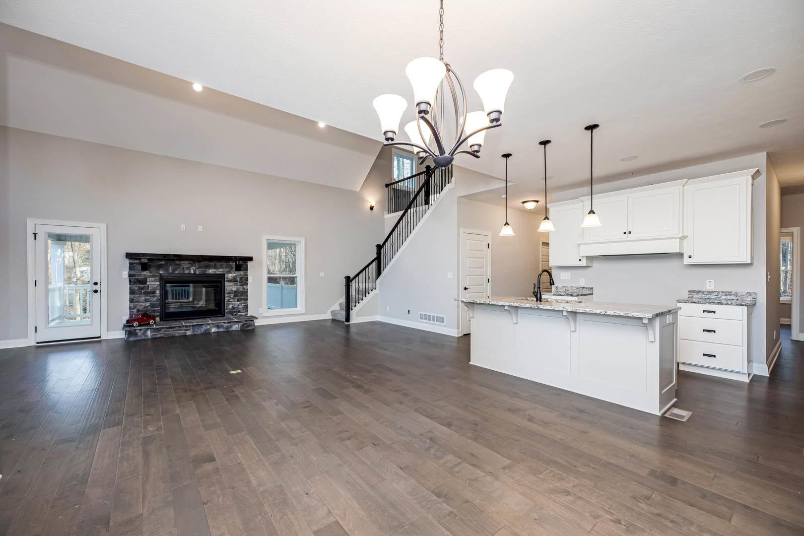 Spacious living room featuring hardwood floors, marble countertop, large chandelier, fireplace with a toy car inside, white cabinetry, and glass-paneled door; windows provide