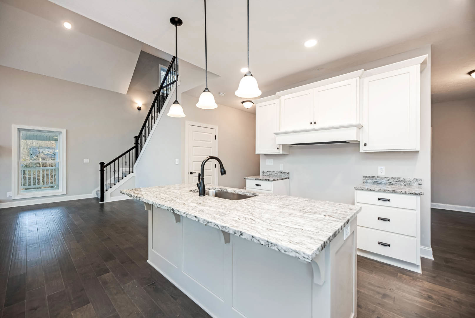 Marble kitchen countertop with built-in sink, white cabinetry with black handles, adjacent staircase with white railing, light wood flooring, white door and frame