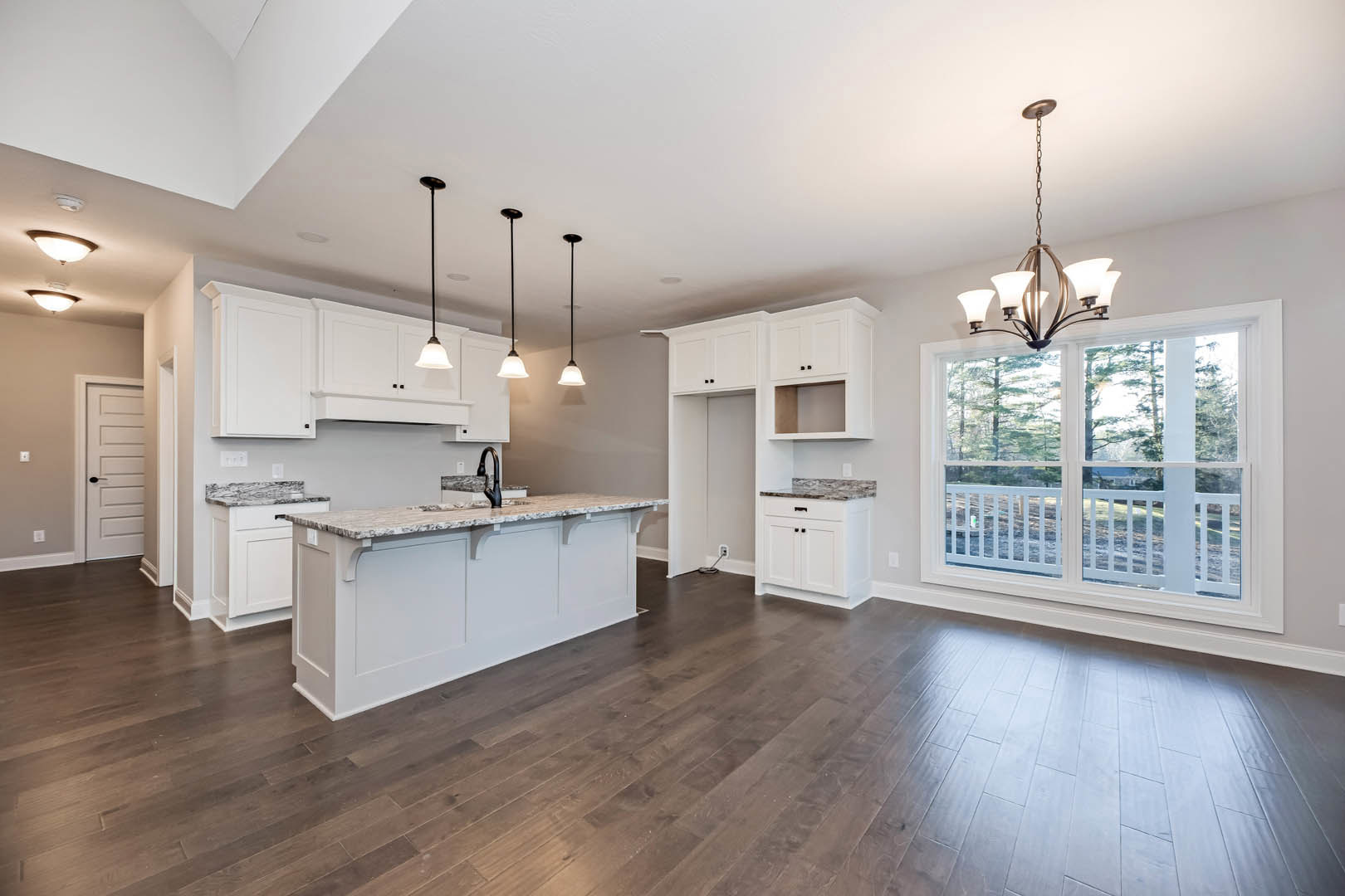 Open kitchen and dining area with dark hardwood flooring, white cabinetry, marble countertop, window with white railing, modern light fixture, and white door with black handle.