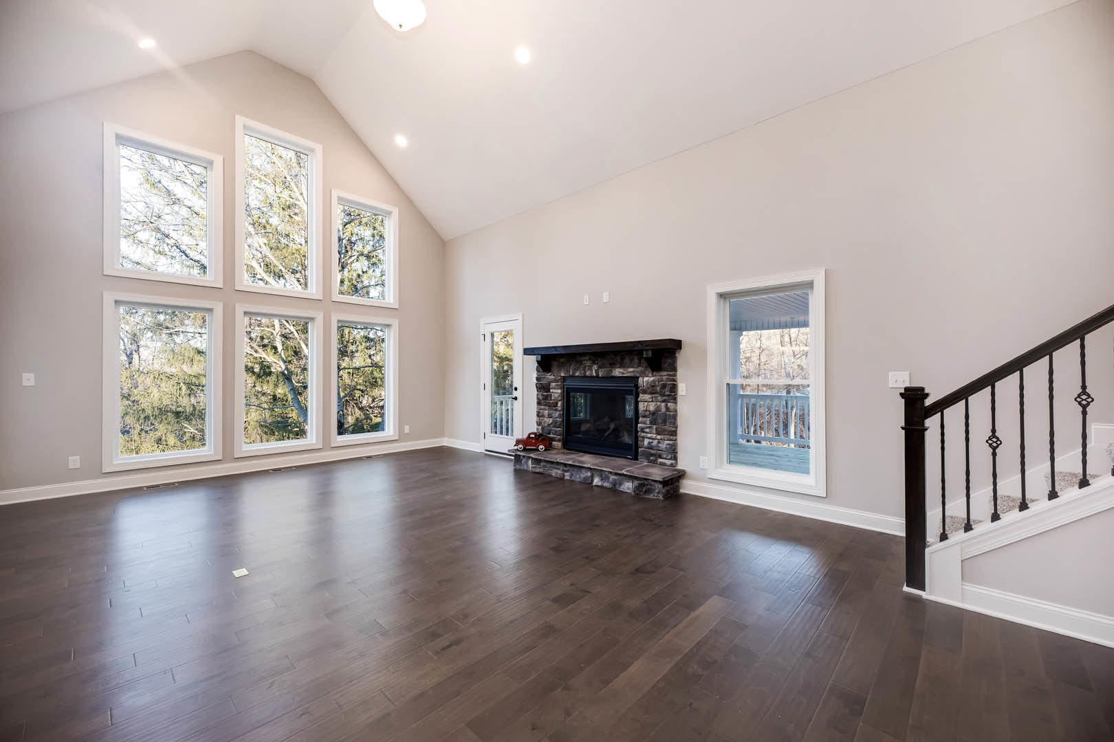 Spacious living area featuring hardwood floors, central stone fireplace, large windows with porch view, and adjacent staircase with wood railing