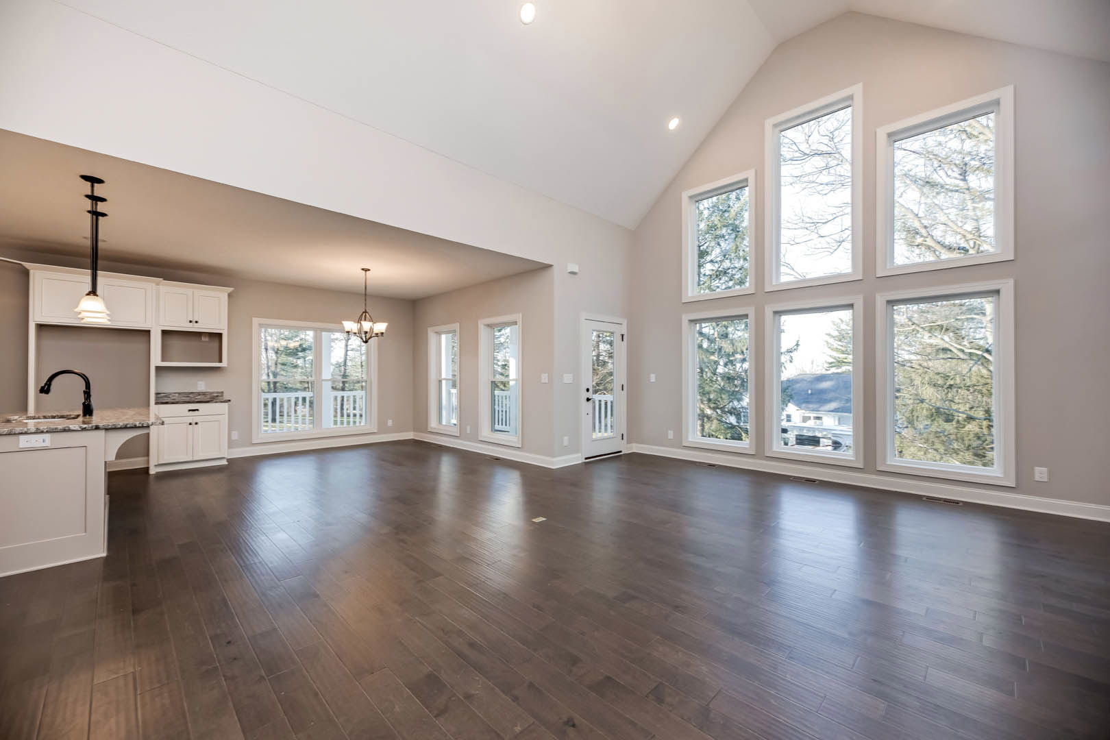 Spacious room featuring hardwood floors, expansive windows with views of leafy trees, and glass doors framed by white walls