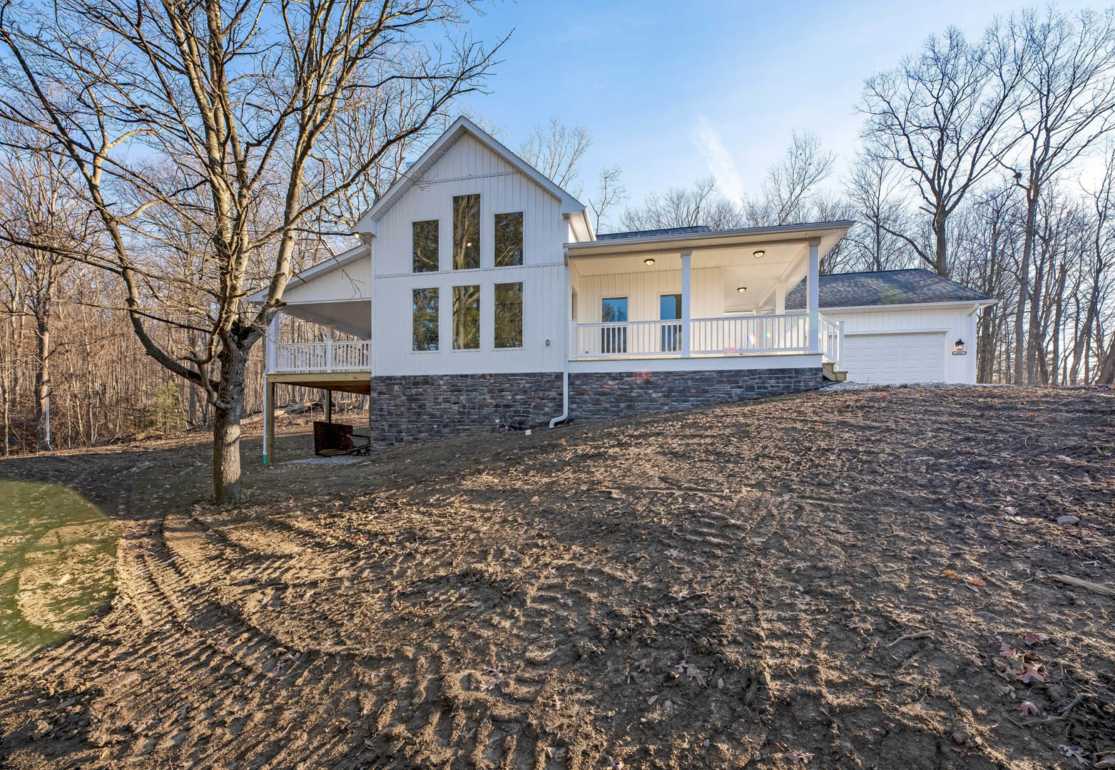 White house with wide covered porch, white railings, large front-facing windows, mature tree beside entry, cloudy sky overhead, dirt ground in foreground