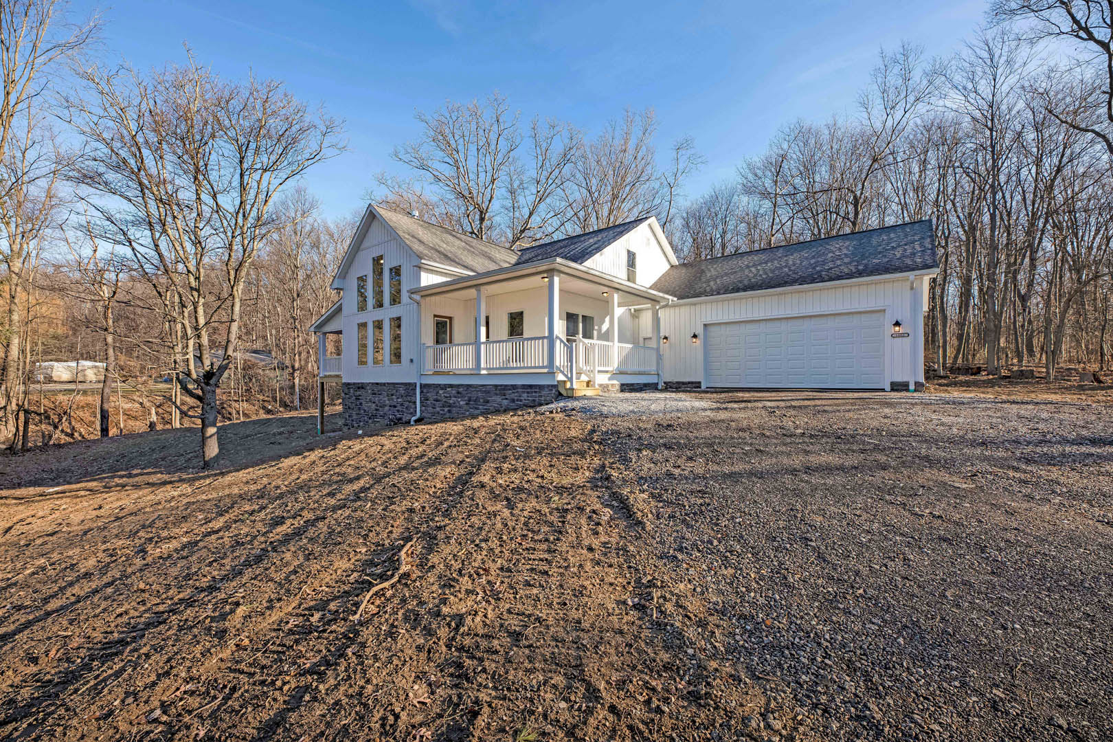 White house with covered front porch, attached garage, bare trees, and dirt driveway under cloudy winter sky