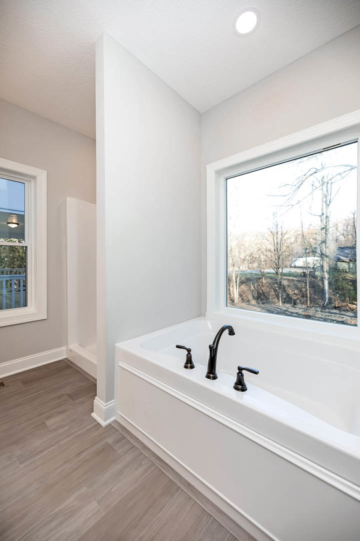 Freestanding bathtub with chrome faucets beneath a large window overlooking trees and a road, white walls, ceiling light, and wood flooring in a modern bathroom.