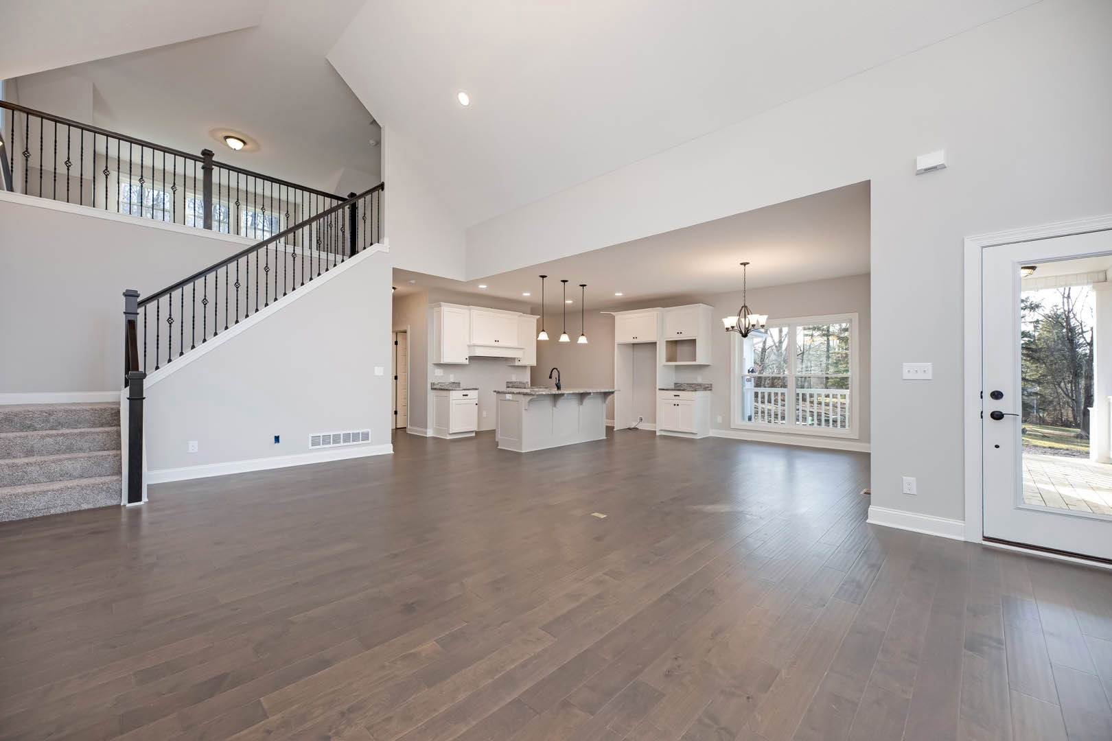 Spacious open floor plan featuring wood flooring, central staircase with white railing, modern kitchen with white cabinets and countertops, large windows offering views of trees