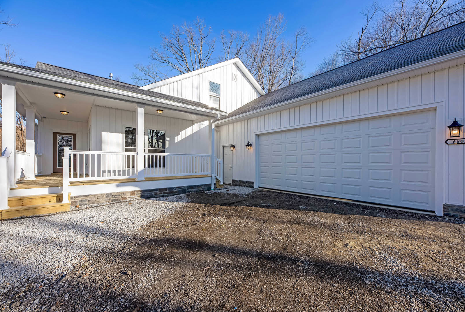White siding house with attached garage, bare trees behind, white railing and vase on front porch, dirt driveway, exterior window close-up.