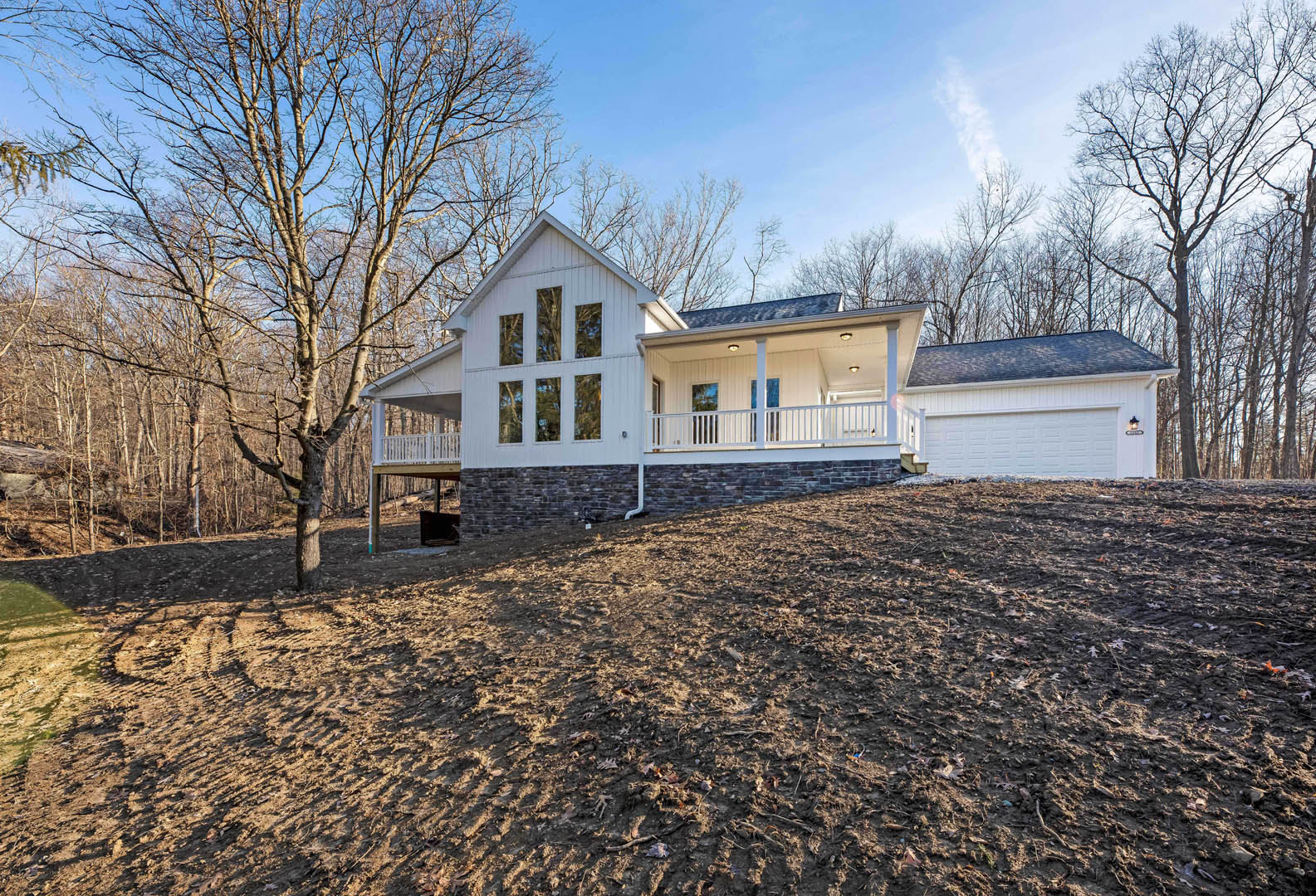 White contemporary house with large windows, situated on a dirt hill, surrounded by mature trees and cloudy sky.