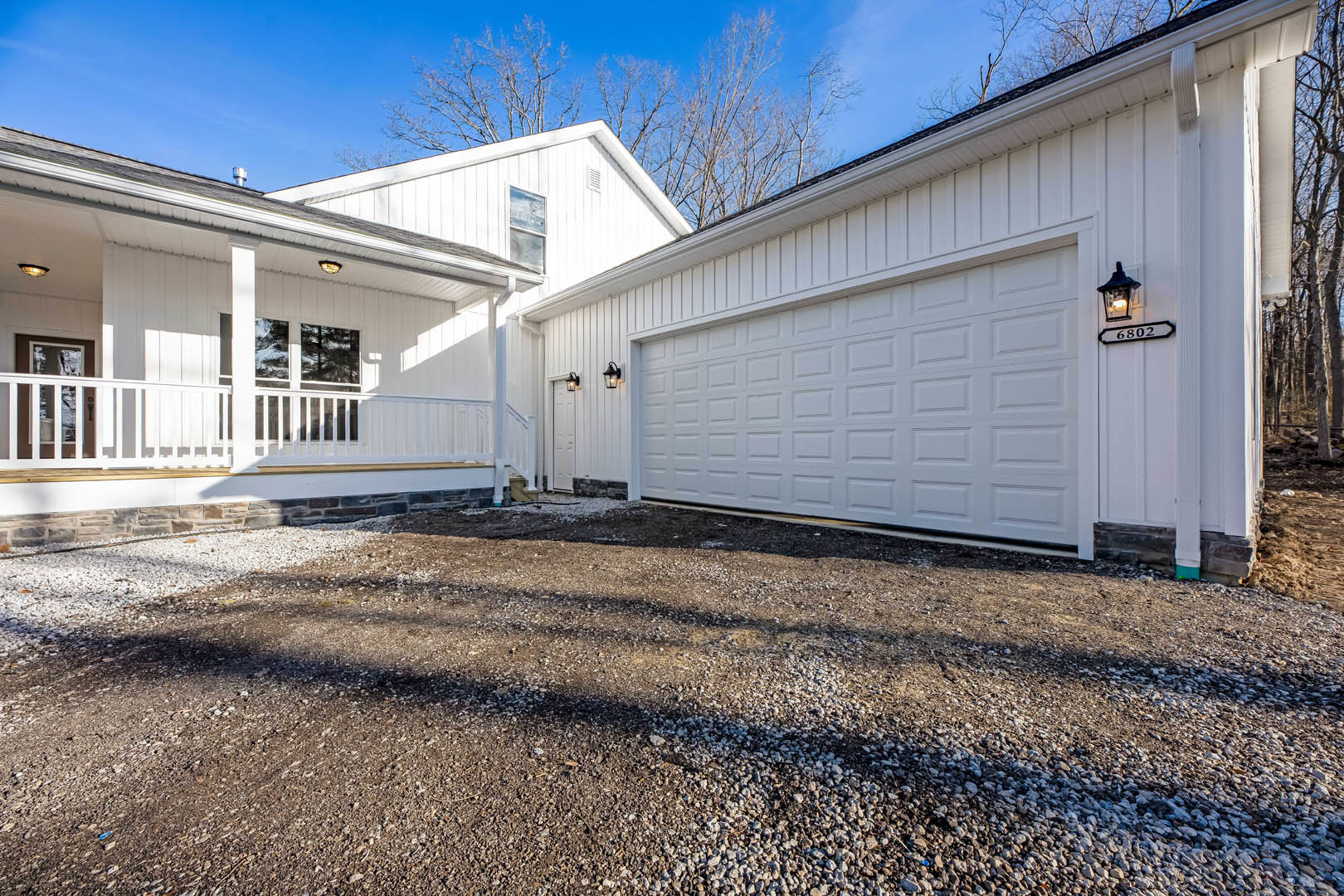 White siding house with attached garage, gravel driveway, white-framed windows, exterior wall lamp, and clear blue sky.