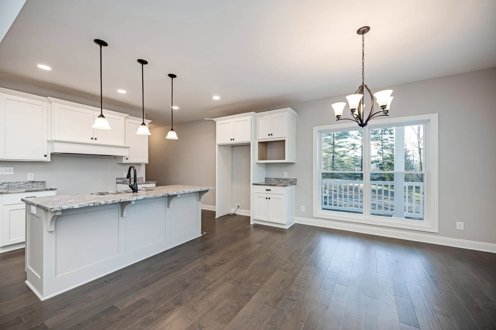 Spacious kitchen featuring a large white island with black faucet, hardwood flooring, white cabinetry, oversized window overlooking trees and porch, and modern pendant lighting