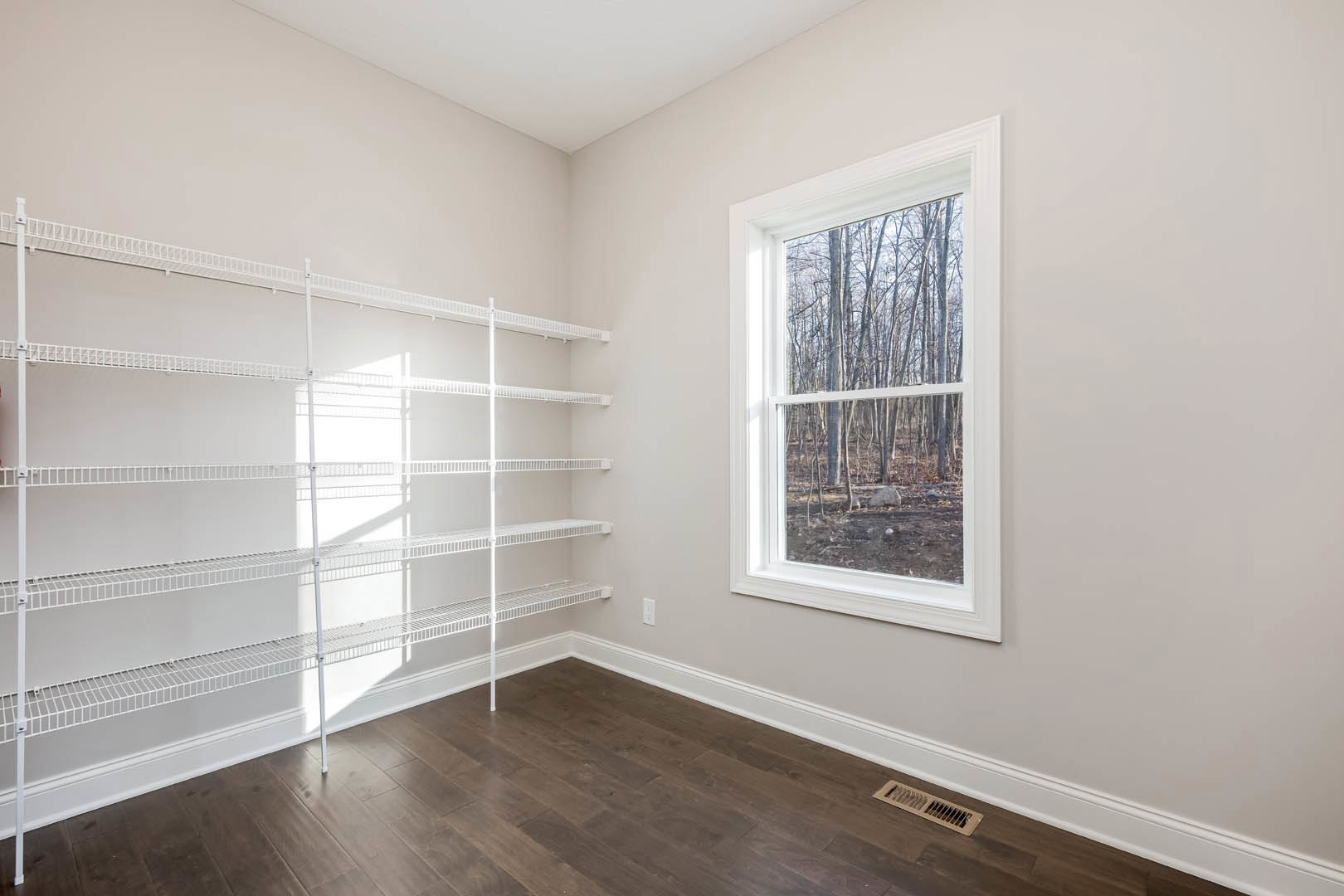 Empty room with built-in white shelves, large window overlooking forest, wood floor with white baseboards, and floor vent