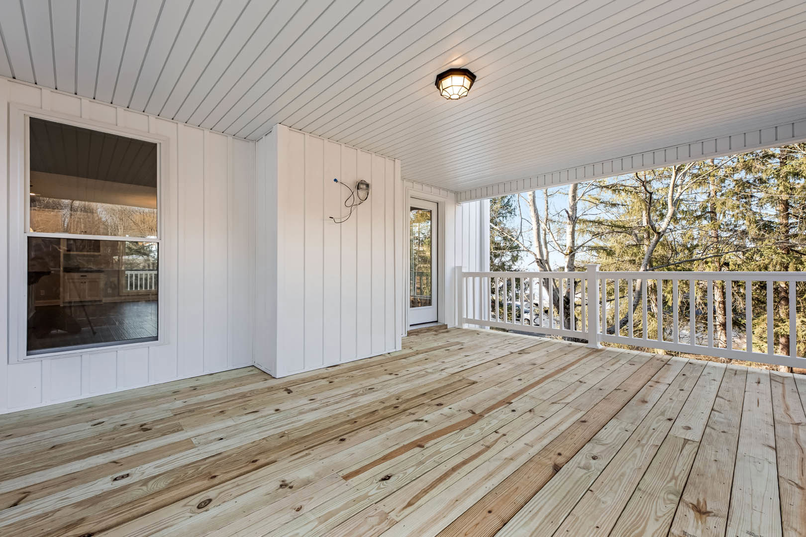 White deck with wooden flooring, white railing, ceiling-mounted light fixture, white door, window overlooking neighboring house, close-up of exposed wire.