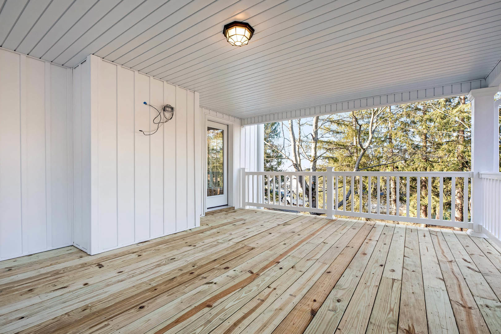 Wooden deck with white railing and white wall, ceiling-mounted light fixture, window overlooking trees, close-up of wire along floor