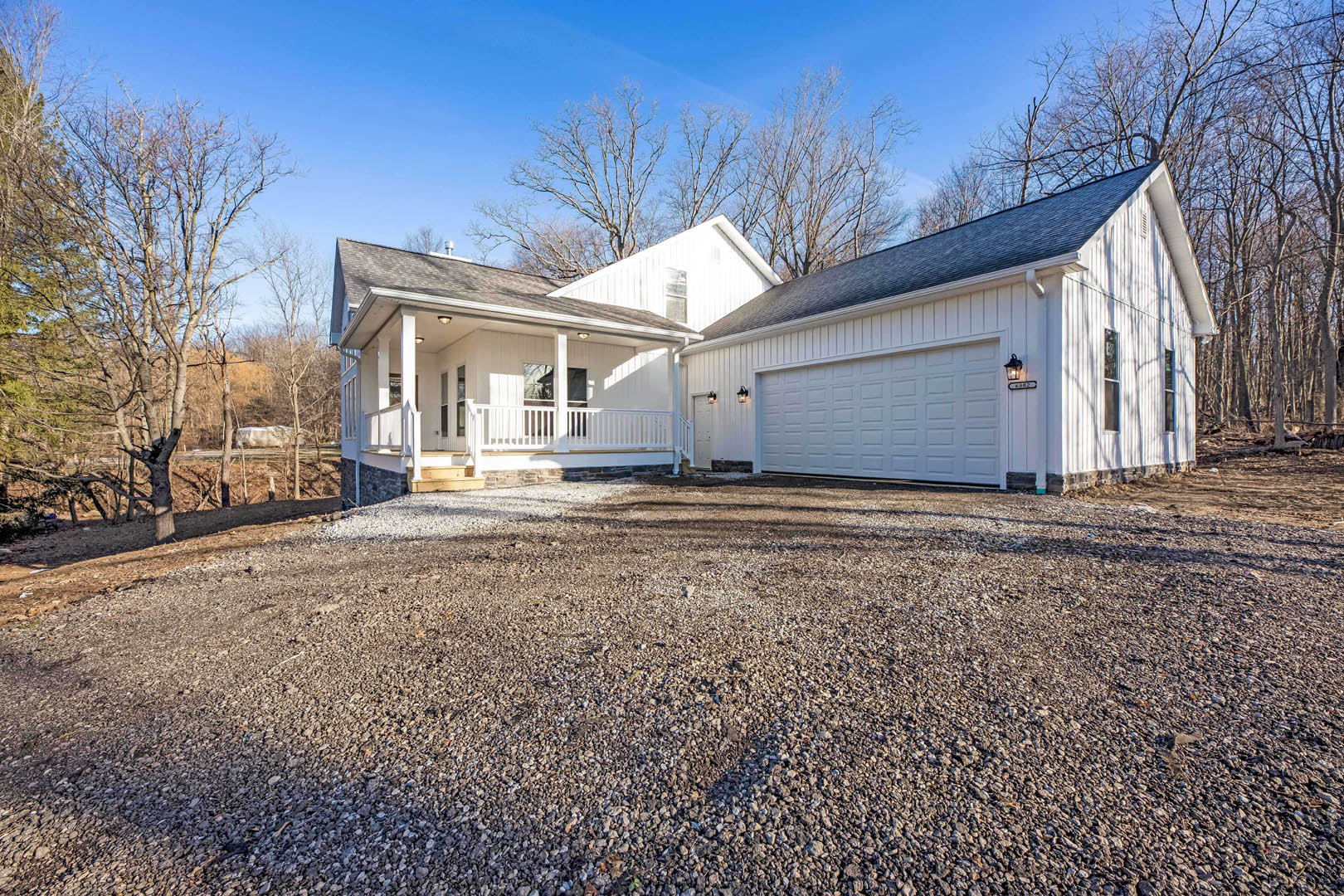 White siding house with attached garage, gravel driveway, porch, exterior light, snow-covered ground, and trees; man standing in front of closed garage door.