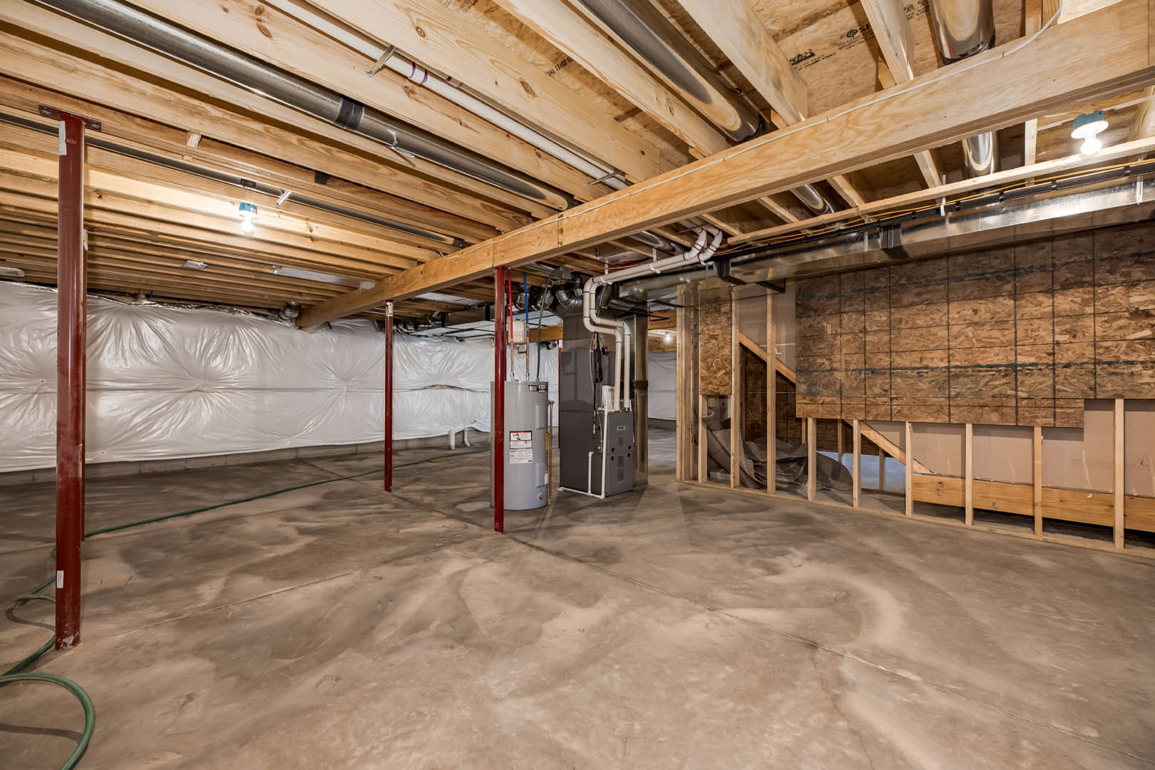 Basement with exposed wooden ceiling beams, large grey tank connected to pipes, concrete floor, white plastic bag hanging from ceiling, and scattered papers on grey surface.