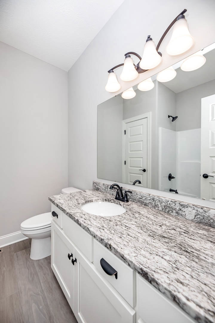 Marble bathroom countertop with undermount sink, black faucet, illuminated mirror above, white toilet, light-colored tile walls, and cabinetry below.