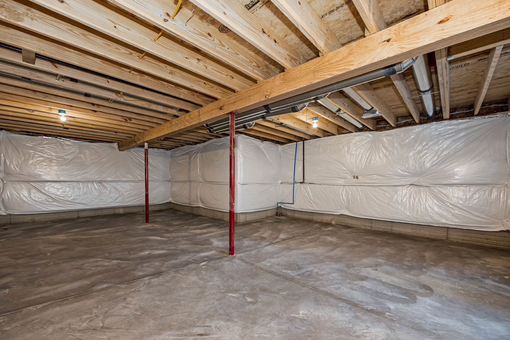 Basement under construction with exposed wooden beams, concrete floor, and walls covered in white plastic sheeting for insulation