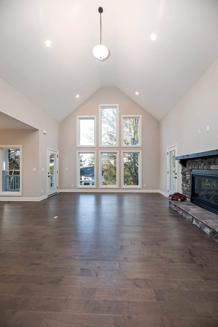 Spacious living room featuring wood flooring, central fireplace with stone surround, large windows offering views of trees, white plaster walls, and ceiling molding.
