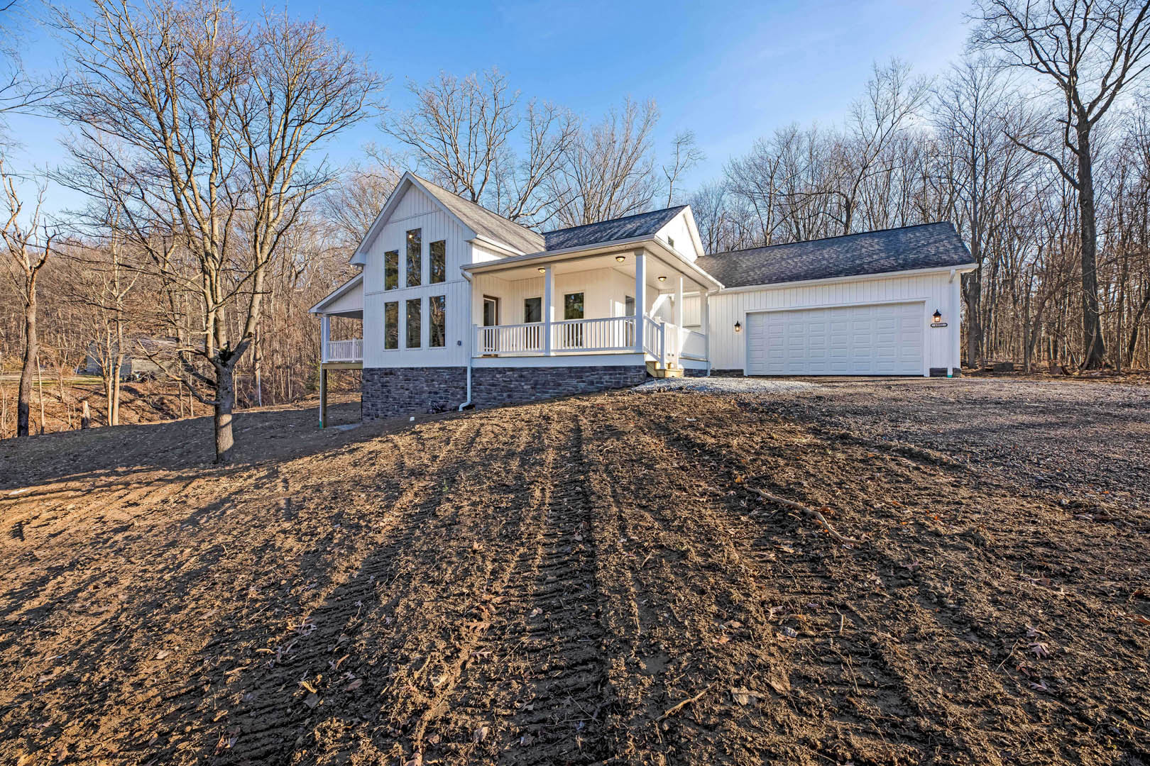 White house with covered porch, black roof, and attached garage, large concrete driveway bordered by winter trees and cloudy sky.