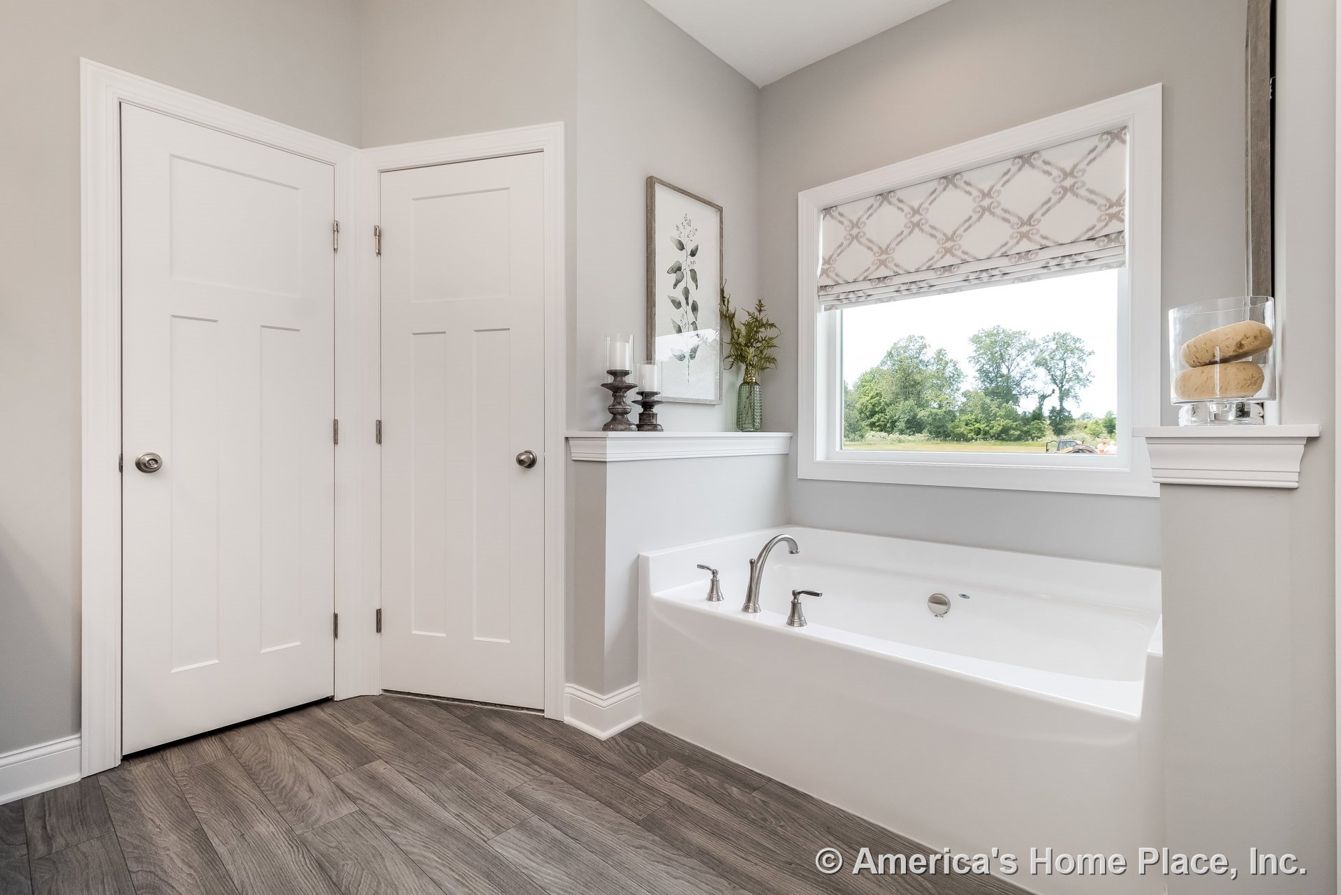 Built-in soaking tub with chrome fixtures set beneath a large window with patterned shade, gray wood-look flooring, white paneled doors, half wall ledge, and light gray painted