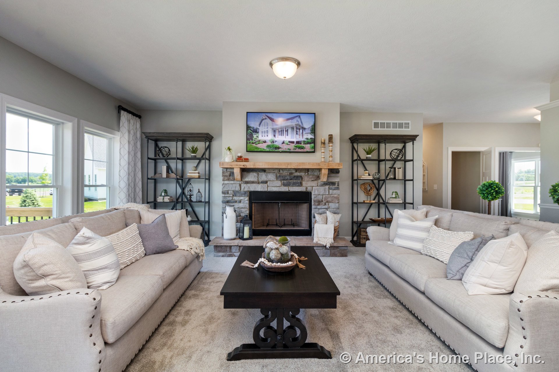 Stone fireplace with wood mantel centered between metal shelving, large windows with white trim, neutral carpet flooring, built-in ceiling light fixture, and open doorway