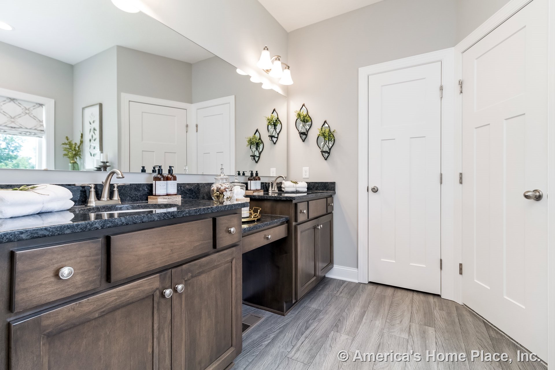 Double sink vanity with dark wood cabinets, granite countertop, large wall mirror, gray tile flooring, built-in overhead lighting, neutral wall color, and double doors with trim.