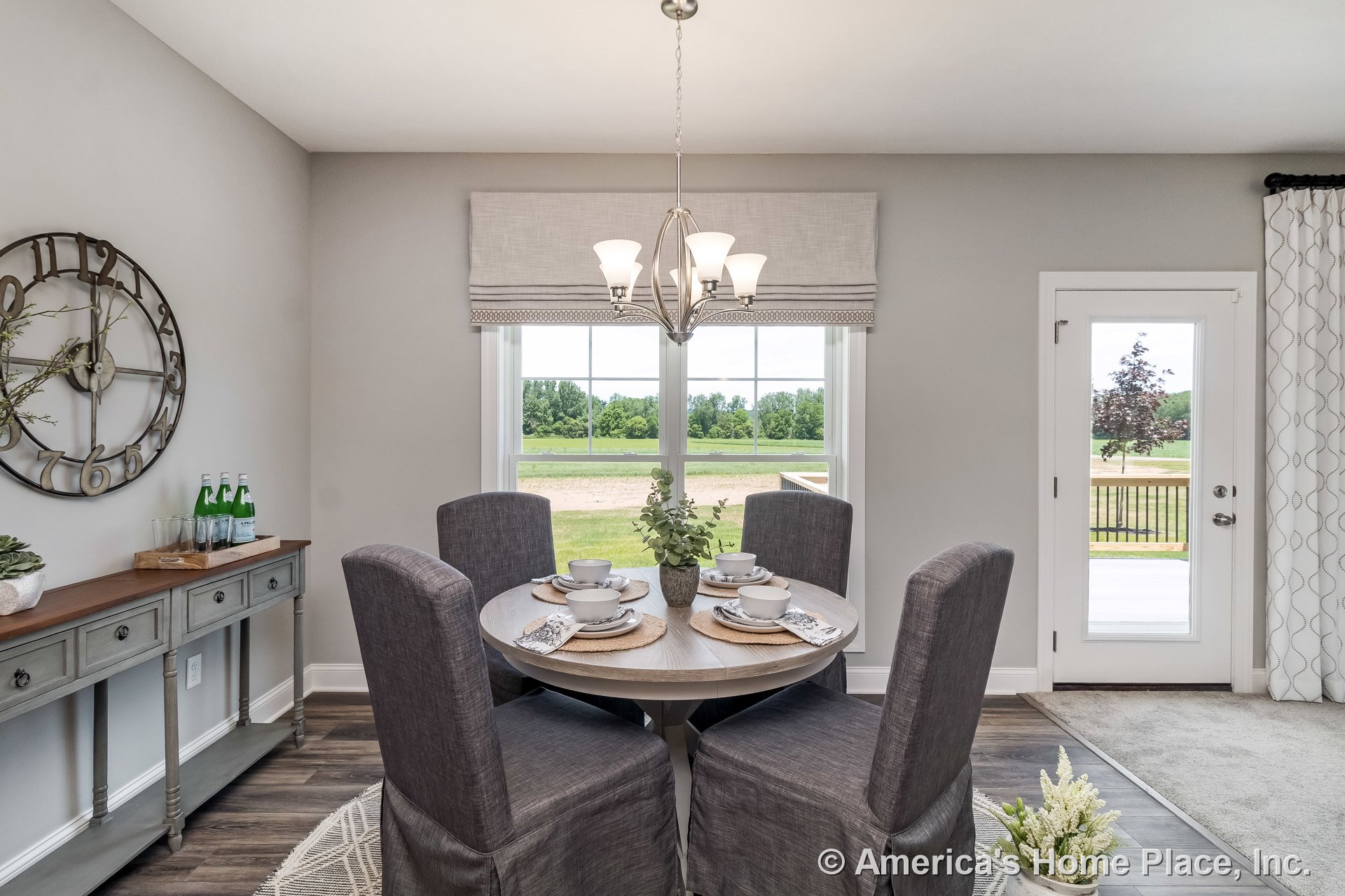 Round wood dining table centered under a brushed nickel chandelier, surrounded by large windows with white trim and a glass door leading to patio; gray console cabinetry along one