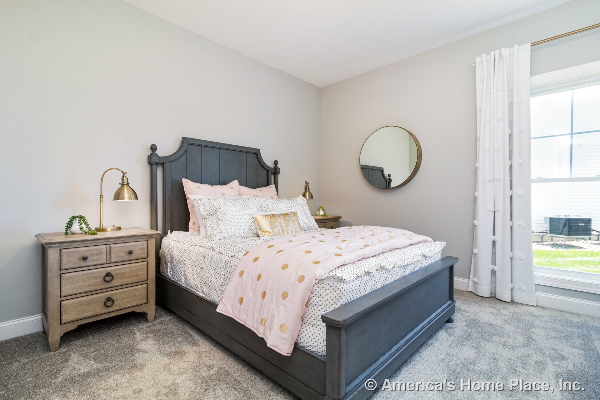 Light gray carpet flooring, neutral painted walls, large window with white trim and white curtains, wood nightstand cabinetry, round wall mirror, white baseboards, built-in ceiling