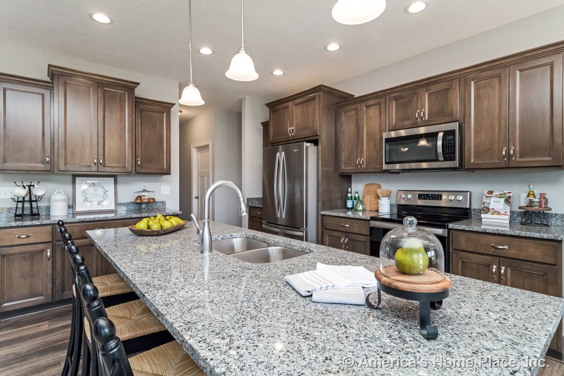 Granite island with undermount sink, dark wood upper and lower cabinets, stainless steel refrigerator, built-in microwave above electric range, pendant and recessed ceiling lights