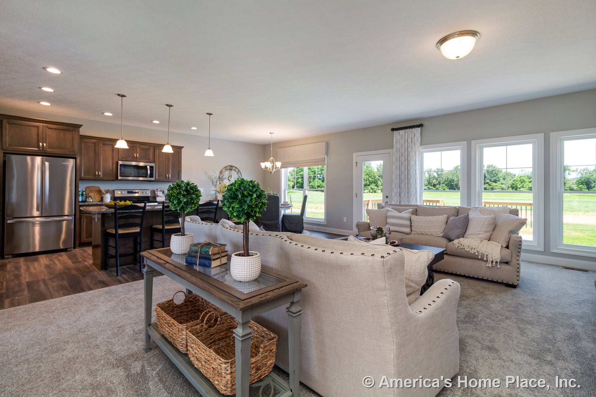 Open-concept living area with carpet flooring, expansive windows framed in white trim, kitchen featuring wood cabinetry, stainless steel appliances, and pendant lights above the