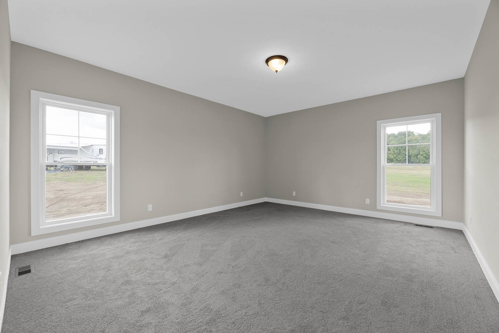 Bright room with large windows, grey carpet flooring, white plaster walls, ceiling light fixture, and views of grass, trees, and a camper outside