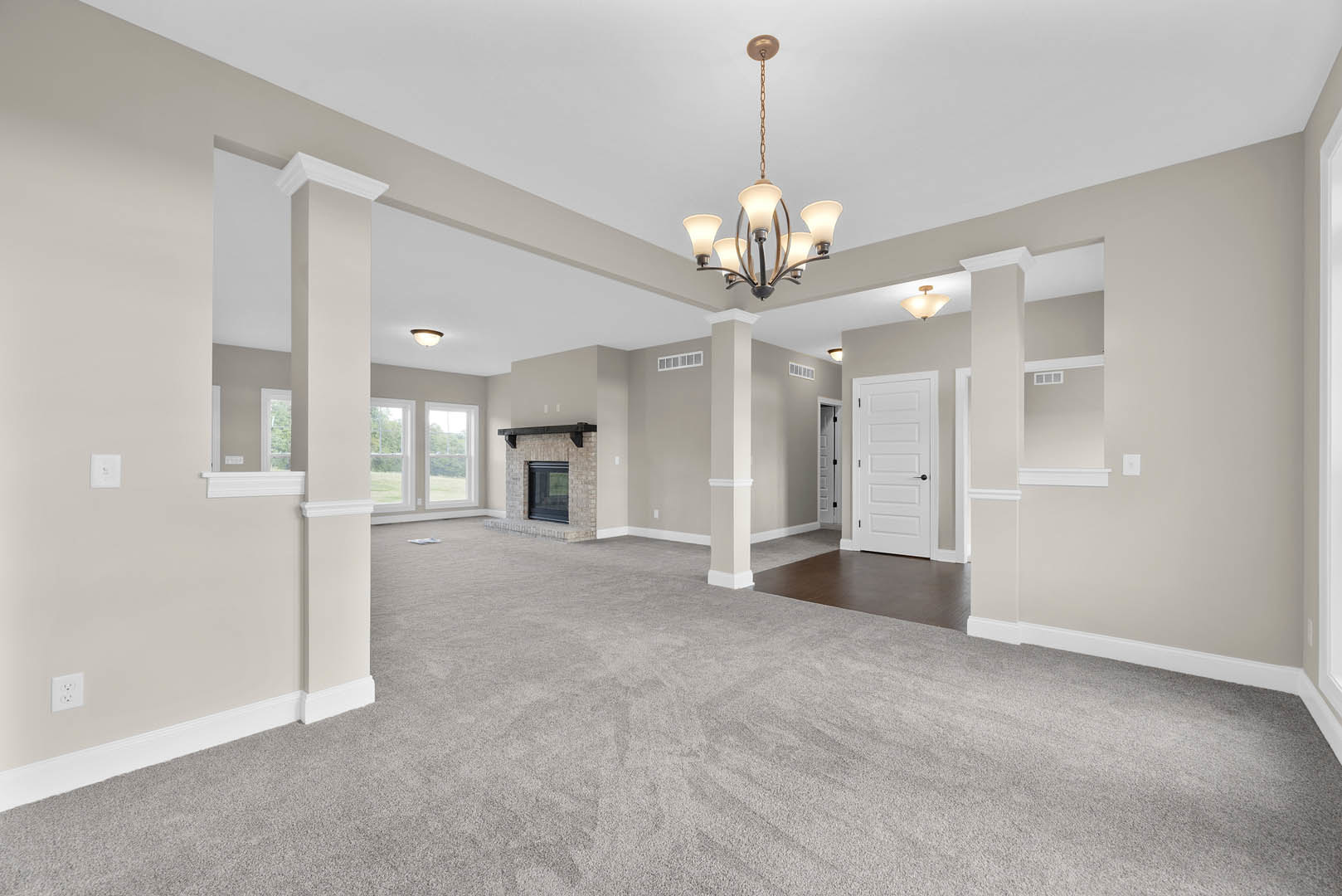 Living room with carpeted floor, white columns, white fireplace with mantel, ornate chandelier hanging from ceiling, white door with black handle, plaster walls with crown molding