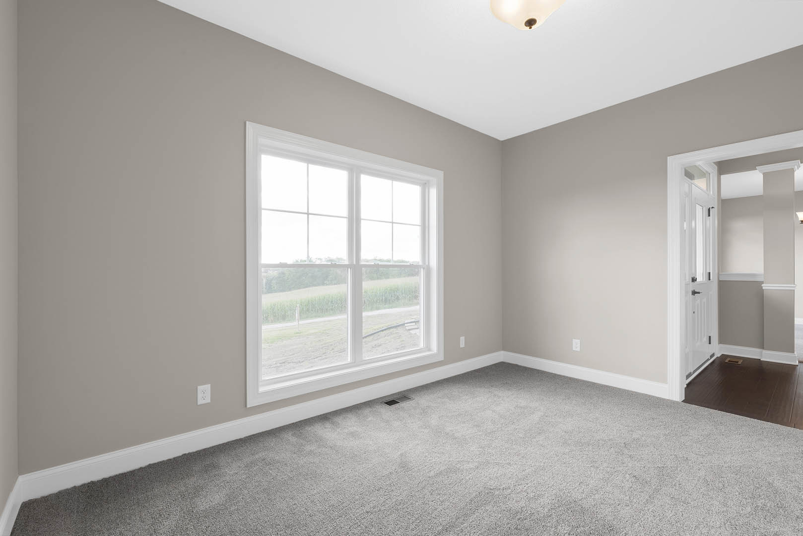 Bright room with white walls, large window overlooking grassy field, soft grey carpet flooring with floor vent, white door in hallway, ceiling light fixture