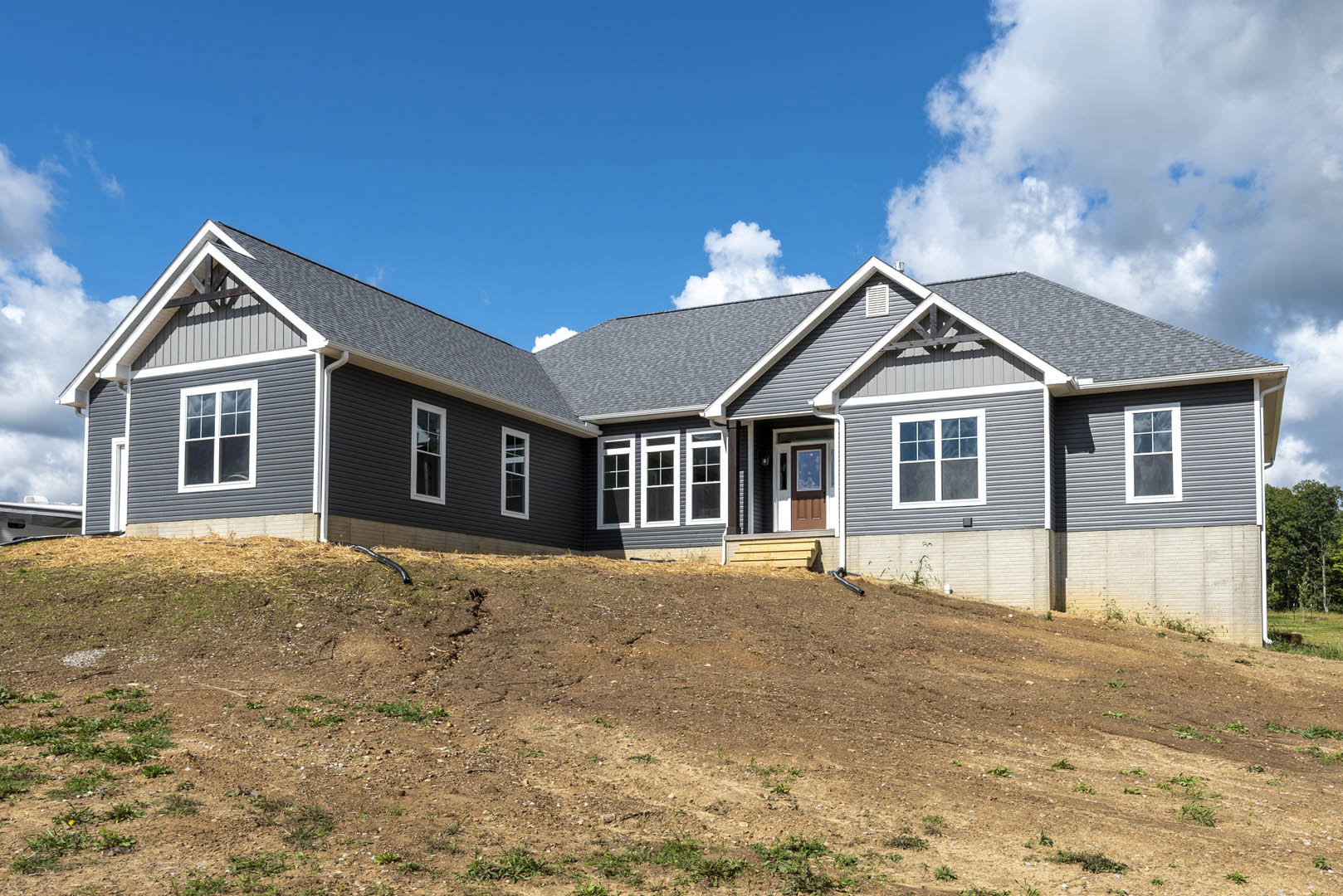 Two-story home with white trim windows and gray roof, situated atop a dirt hill under a partly cloudy sky