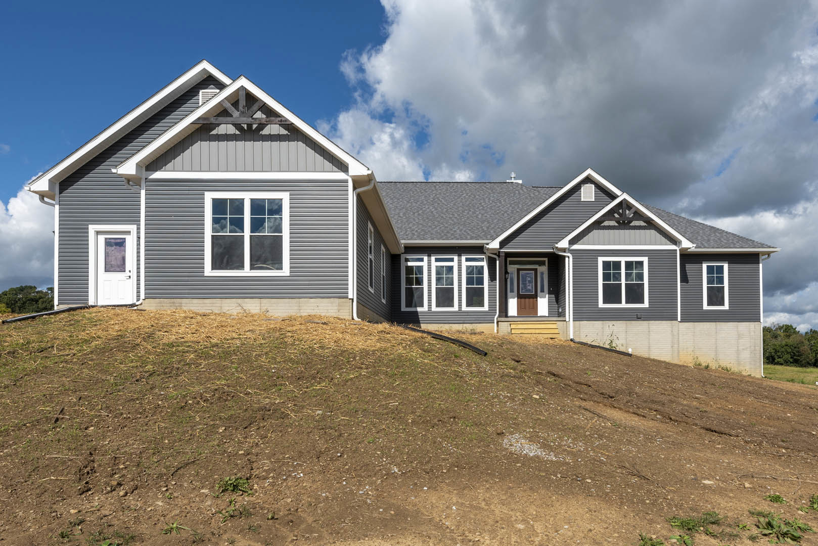 White siding house with white-framed windows and door, situated on a dirt hill with a black pipe, under a grey cloudy sky