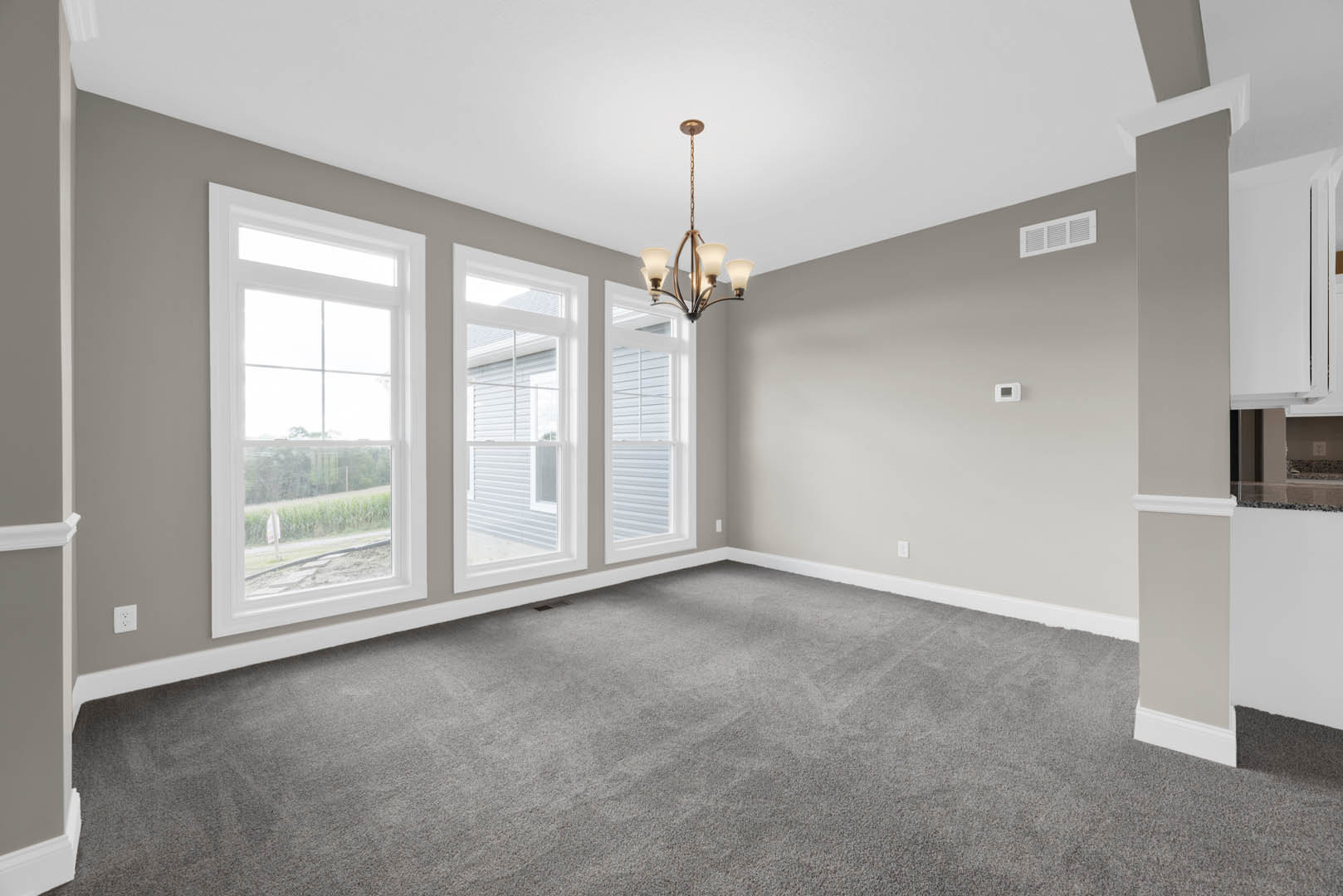 Spacious room with grey carpet, multiple windows overlooking a grassy field, white walls with crown molding, and a modern chandelier hanging from the ceiling