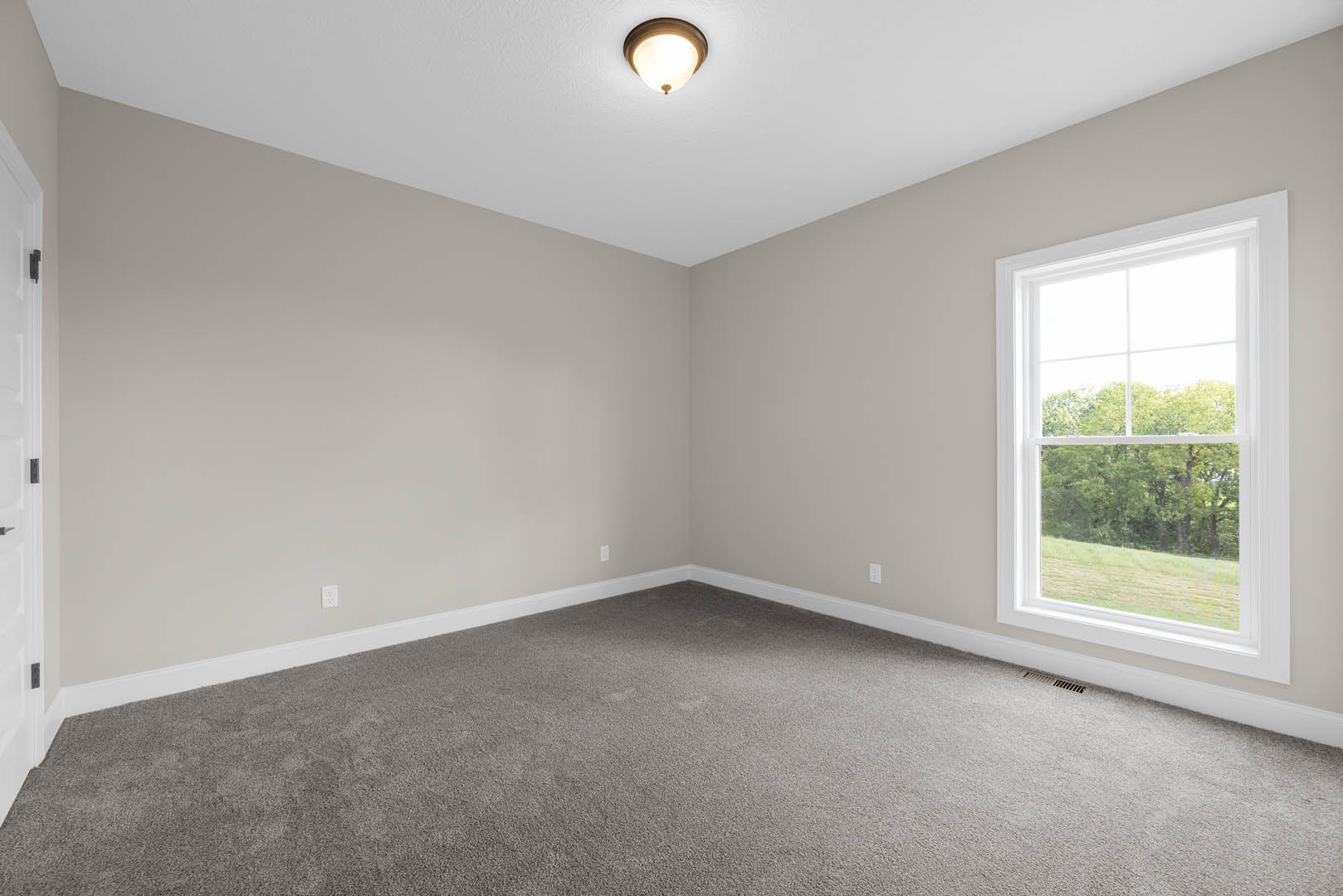 Bedroom with grey carpet, white walls, large window overlooking trees, ceiling light fixture, door with visible hinge, baseboard molding