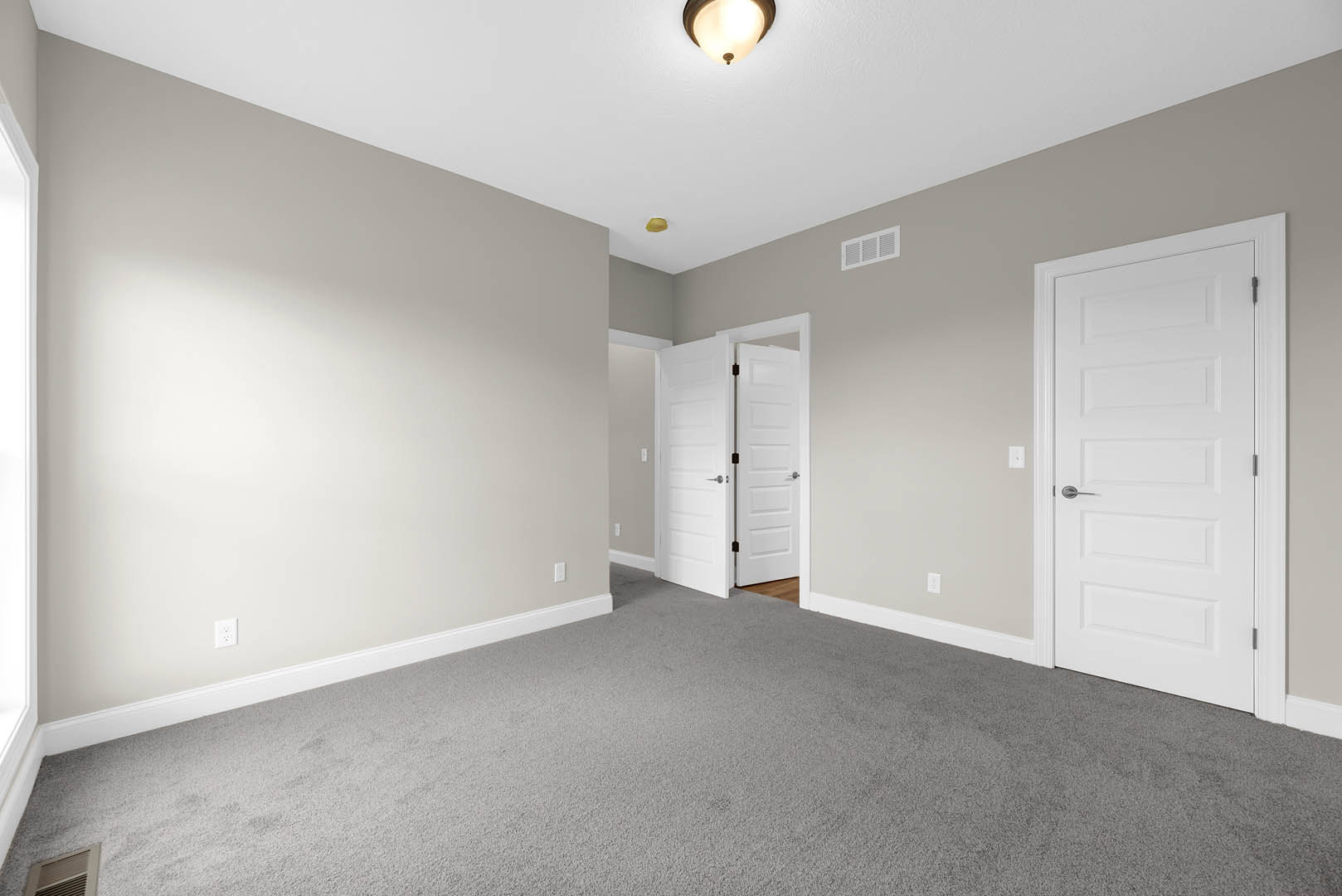 White-walled room with multiple white doors featuring silver handles, light gray carpet flooring, ceiling light fixture, and simple molding along the ceiling and baseboards