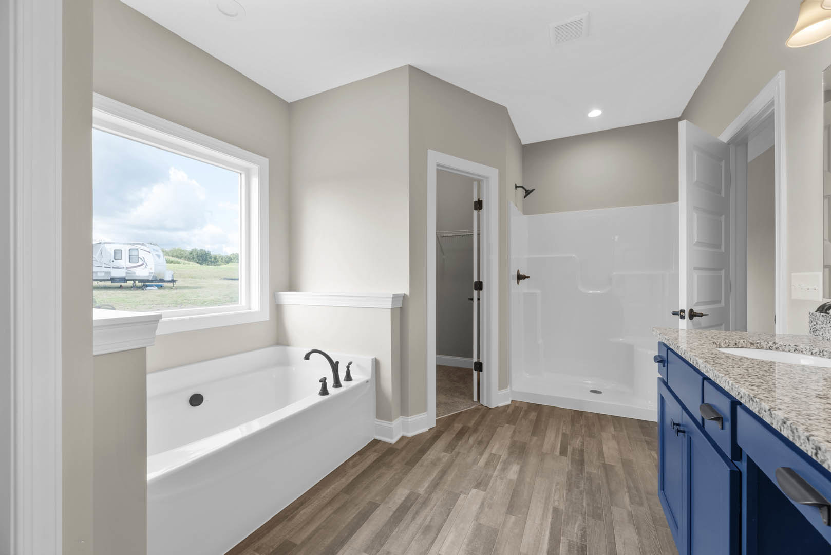Bathroom featuring freestanding bathtub with chrome faucets, glass-enclosed shower, wood flooring, white tile walls, and modern vanity with stone countertop.