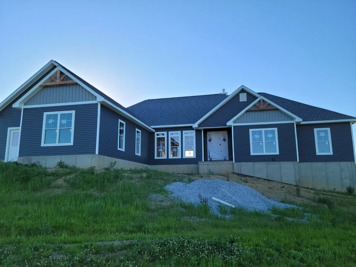 Two-story blue siding house with white trim and multiple windows, situated on a grassy hill under a clear sky