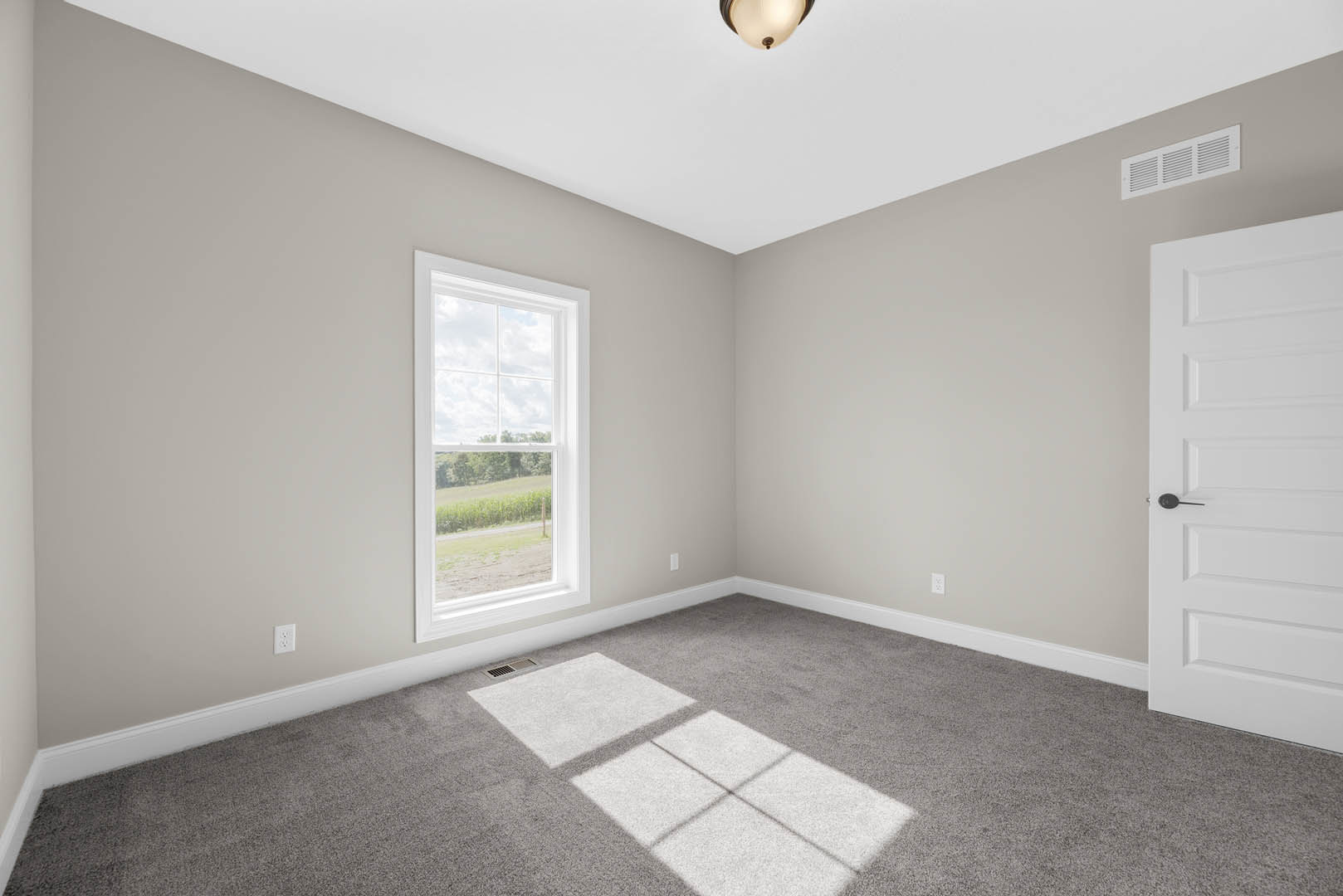 Bedroom with grey carpet flooring, white rectangular rug, large window overlooking a grassy field, white door with black handle, wall vent, and white baseboard molding