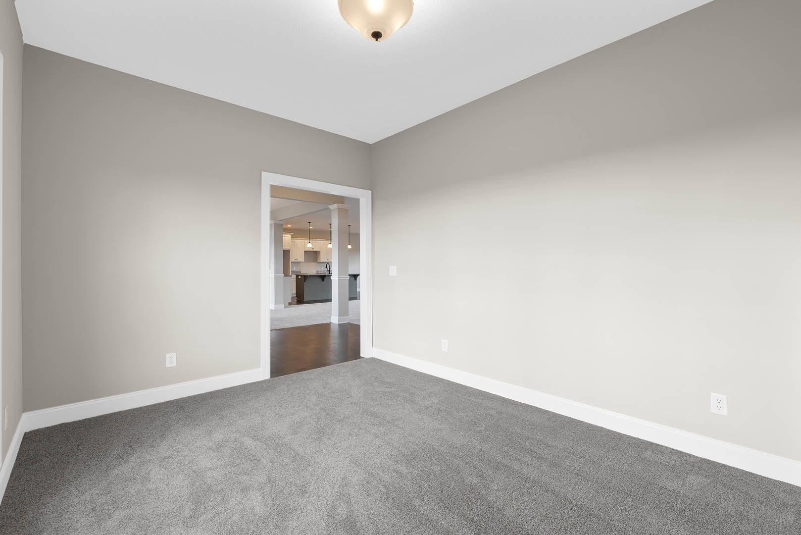 Open doorway leading from a room with brown wood flooring and white walls, kitchen and dining area visible with white pillars, ceiling light fixture, and grey carpet in foreground