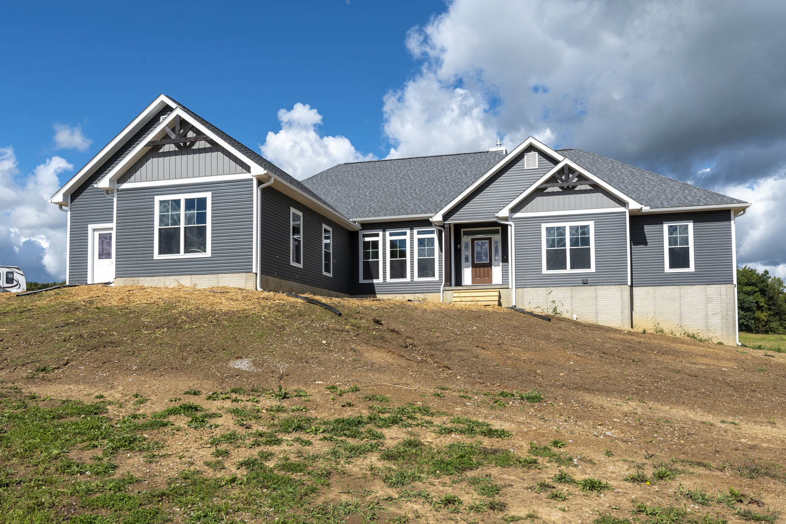 Modern cottage-style home with white-framed windows, situated atop a grassy dirt hill under a blue sky, with a visible entry door and trailer nearby.
