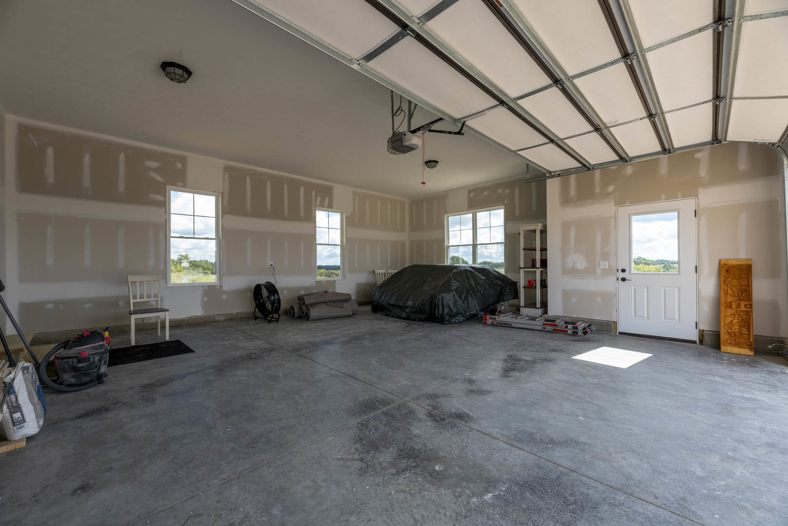 Living room with a black tarp draped over a couch, white chair with black seat, white door with glass window, large window showing trees and sky, paper money on the floor